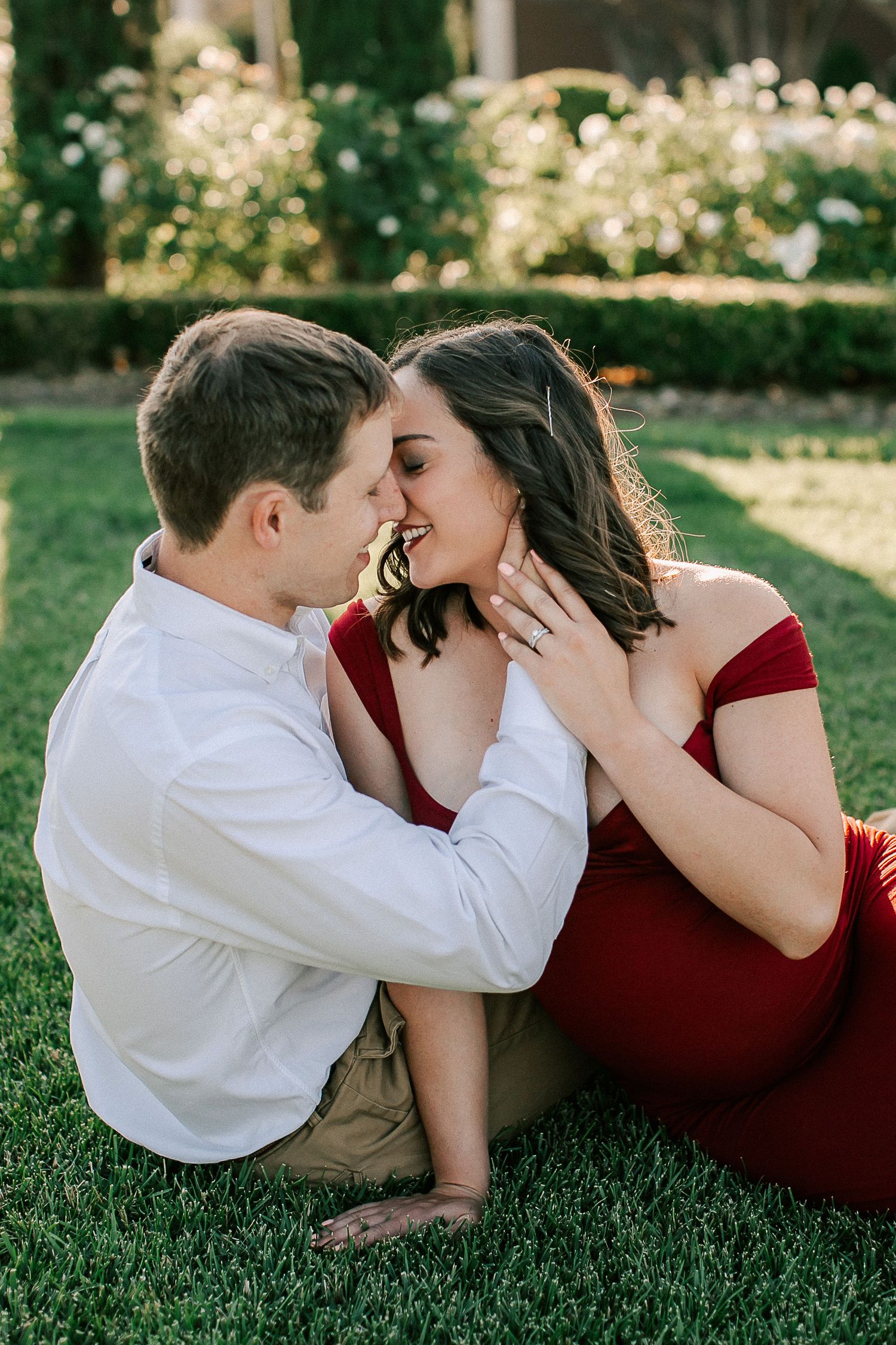 Couple sharing a romantic moment on green grass, woman in red off-shoulder dress, man in white shirt, engagement ring visible.