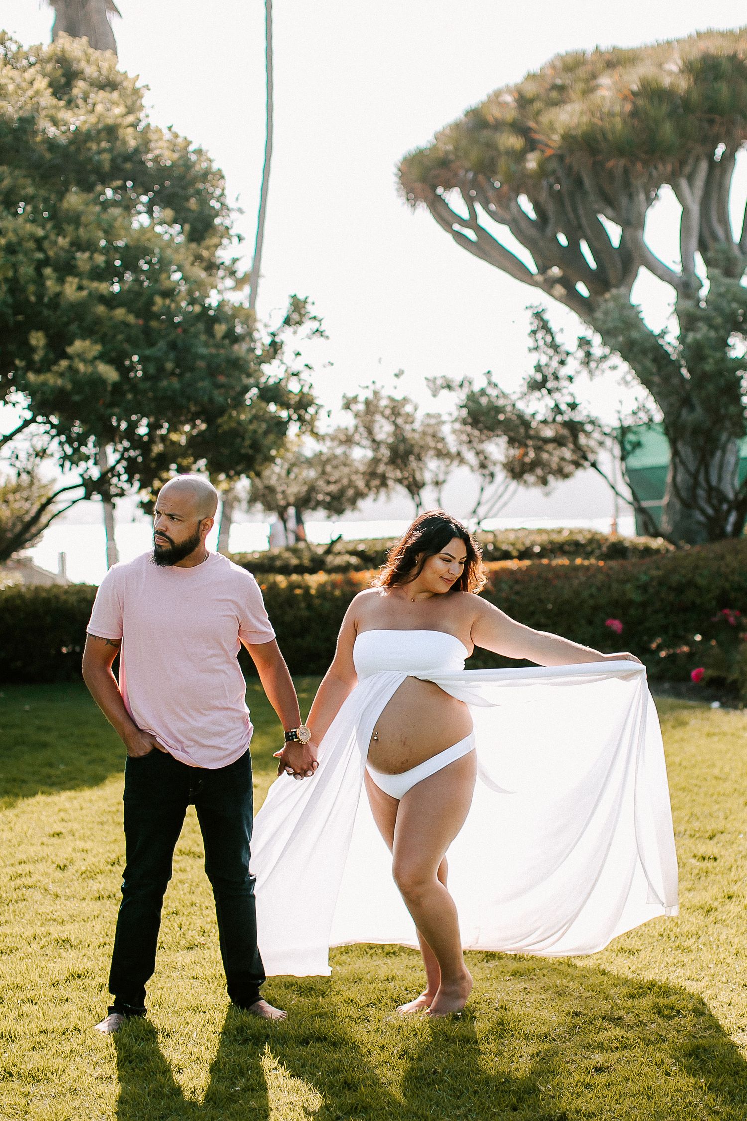 Couple holding hands on green lawn, pregnant woman in white maternity outfit with flowing skirt.