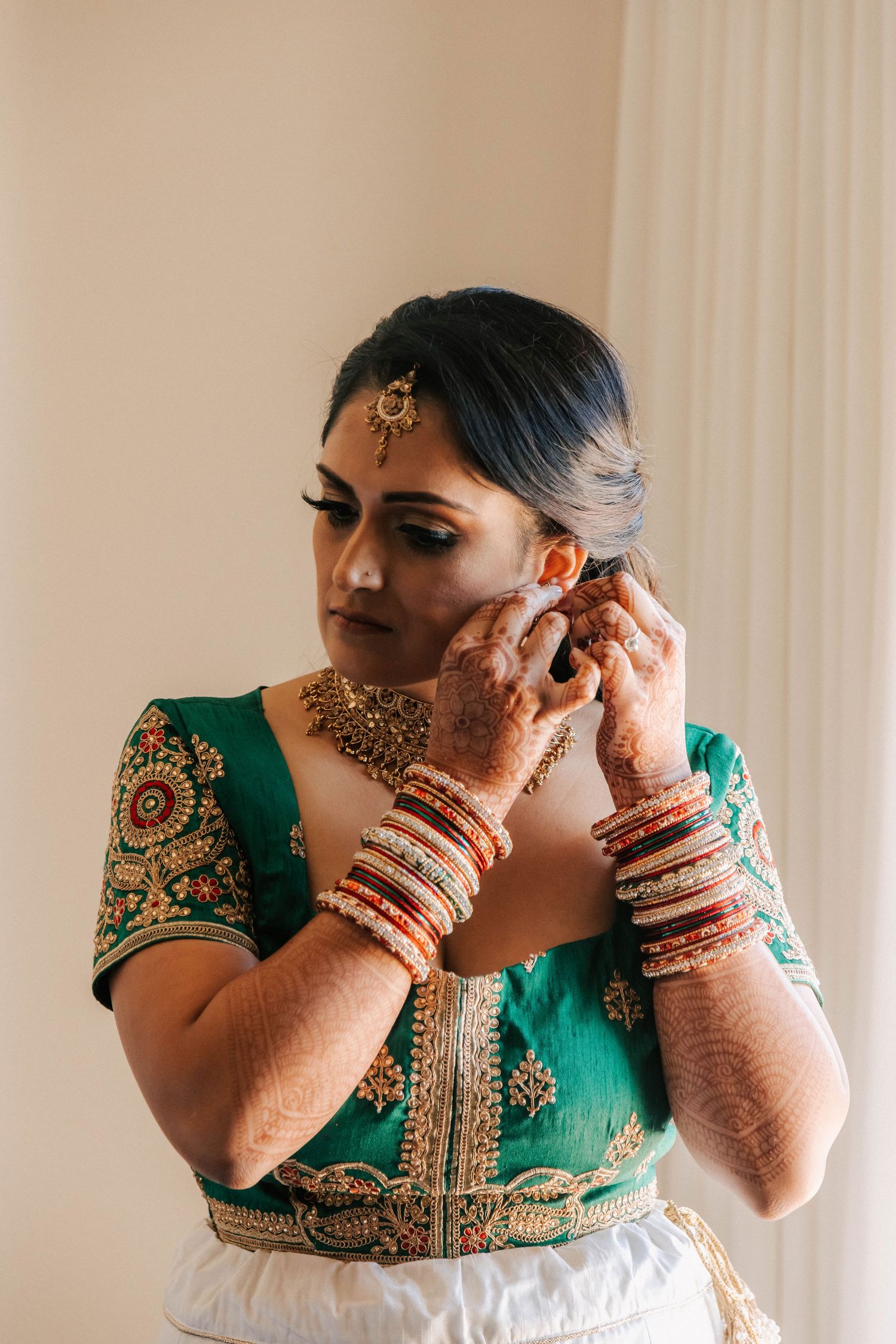 Indian bride in green embroidered blouse adjusts earring while wearing traditional gold jewelry and bangles.