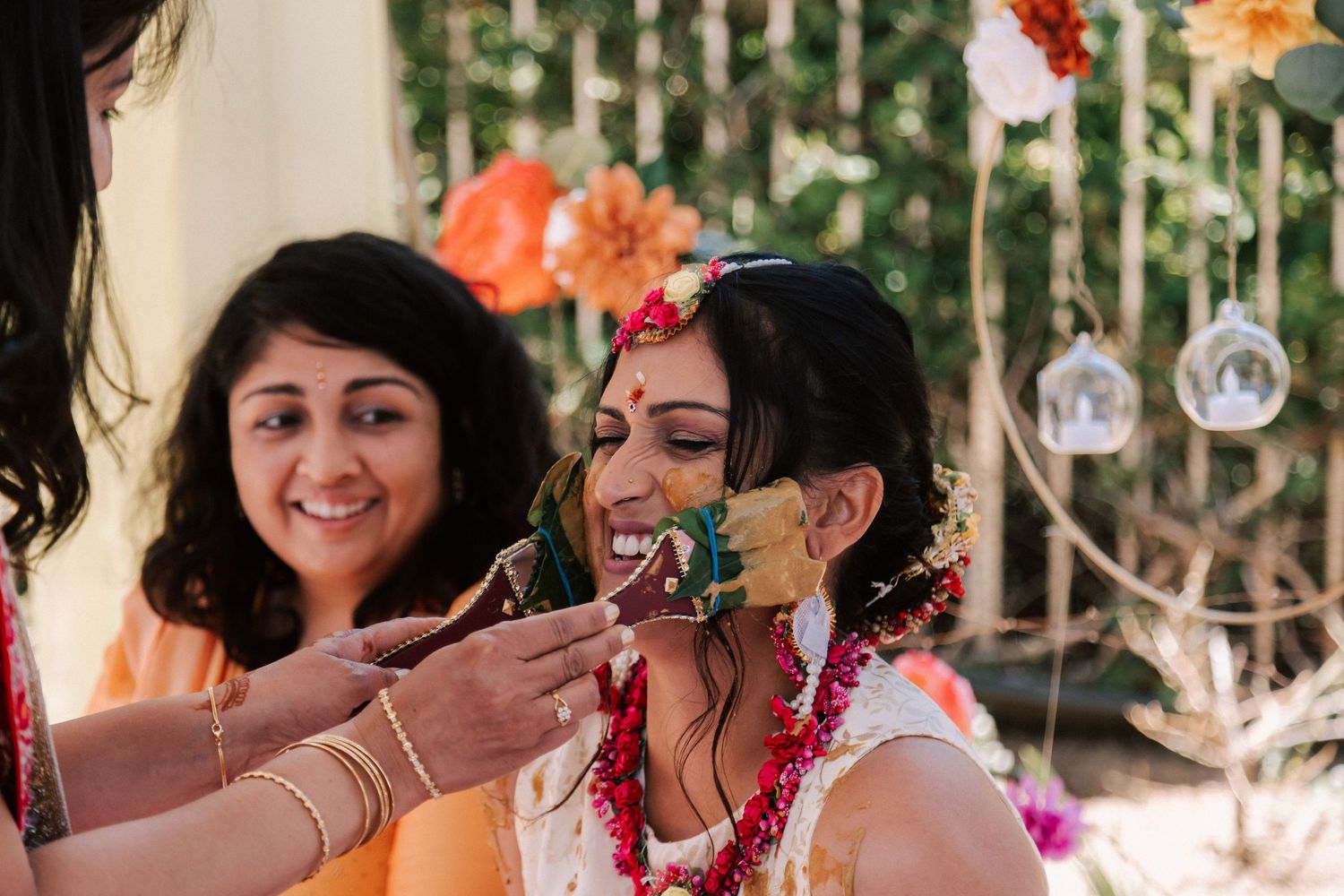 Bride laughing joyfully as attendants apply haldi turmeric paste during Indian pre-wedding ritual.