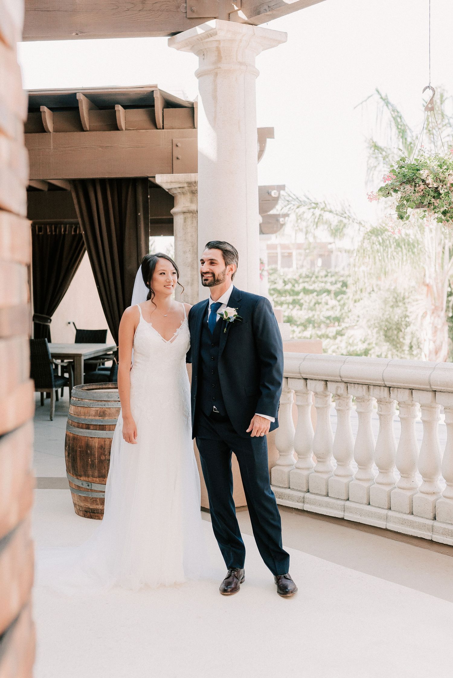 Bride in white gown and groom in navy suit stand together on elegant venue balcony with stone columns.