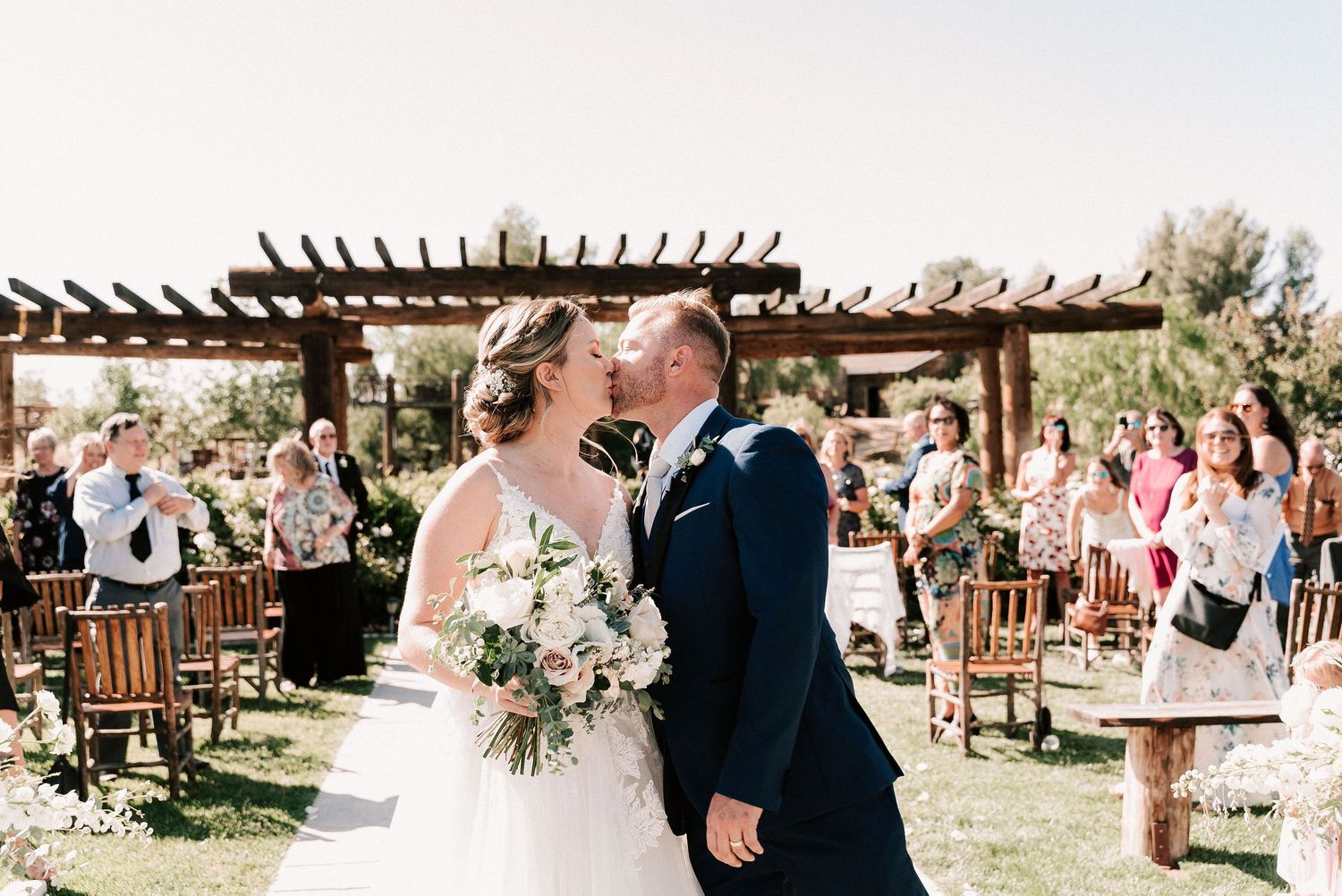 Newlywed couple sharing first kiss at outdoor wedding ceremony with guests cheering and rustic pergola backdrop.