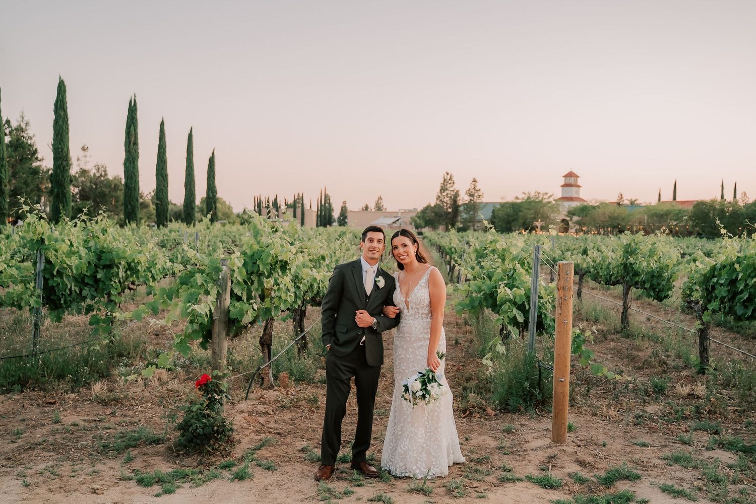 Bride and groom pose together in a lush vineyard at sunset, surrounded by cypress trees and green vines.