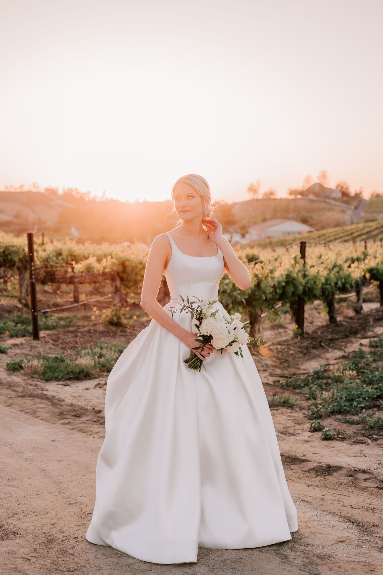 Bride in white ball gown holding bouquet at golden hour in vineyard.