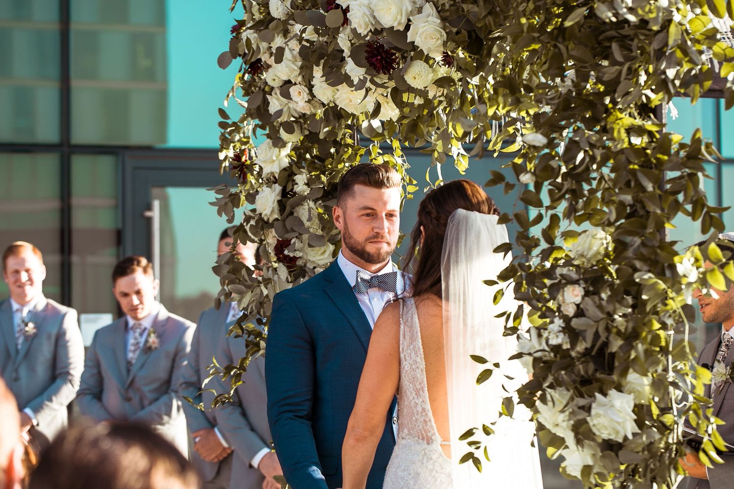 Groom in blue suit smiles at bride under floral arch with white and burgundy flowers at outdoor wedding ceremony.