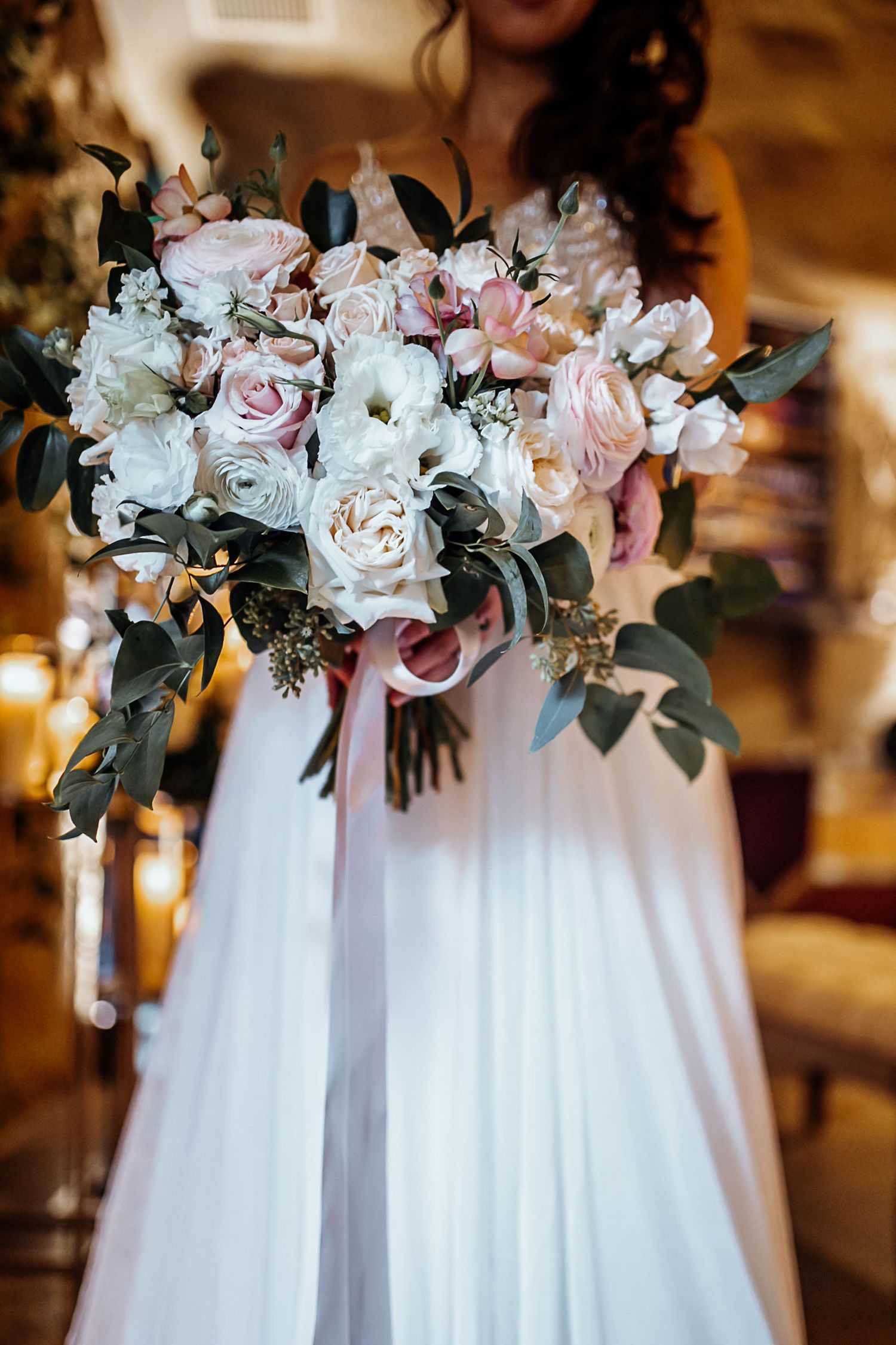 Bride in white gown holding lush wedding bouquet of blush pink and white roses with eucalyptus and pink ribbons.