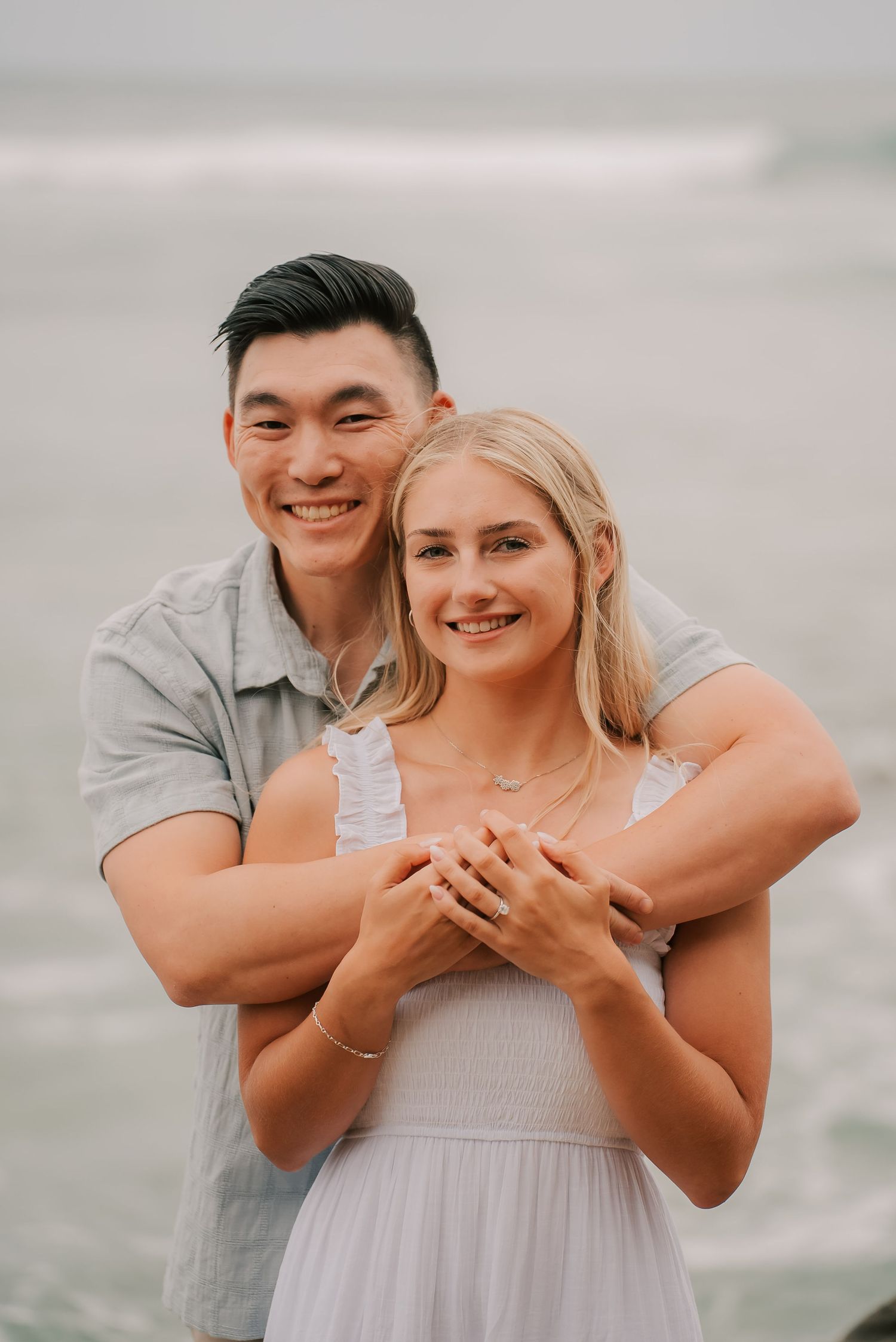Happy couple embracing on beach, man hugging woman from behind, both smiling, ocean waves in background.