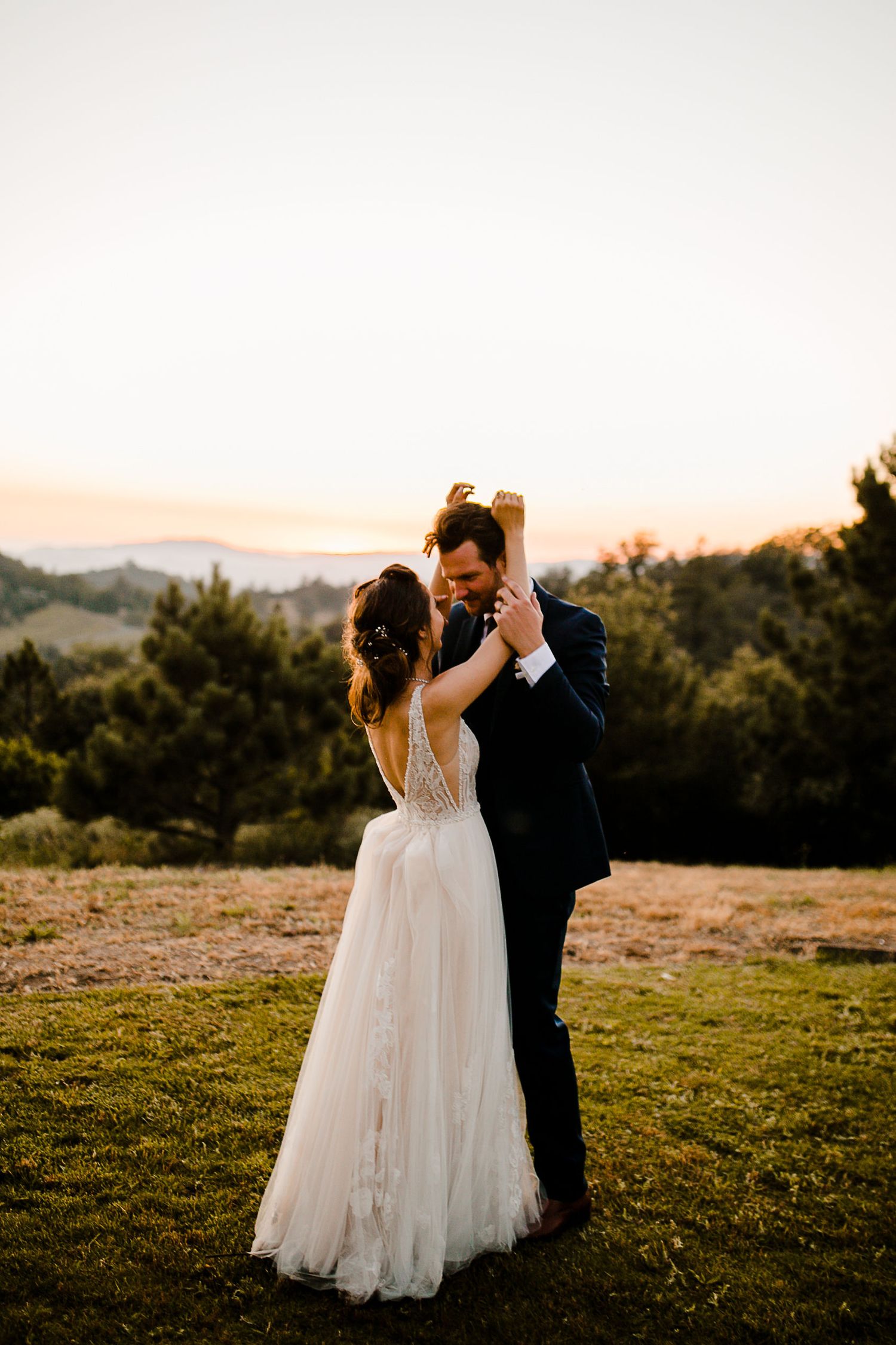 Bride and groom share first dance at golden hour outdoors, surrounded by lush trees and rolling hills at sunset.
