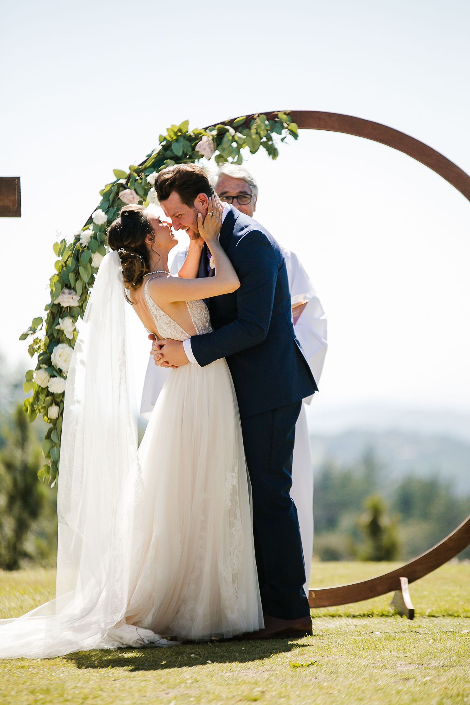 Bride and groom share first kiss under circular wooden arch adorned with greenery and white flowers at outdoor wedding.