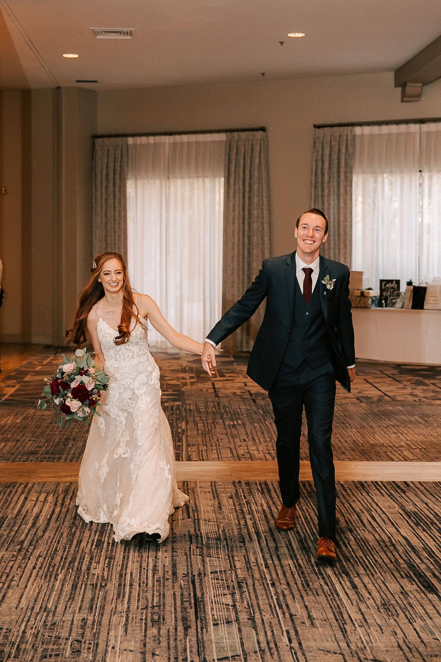 Bride in lace gown holding bouquet and groom in navy suit hold hands while making grand entrance at wedding reception.