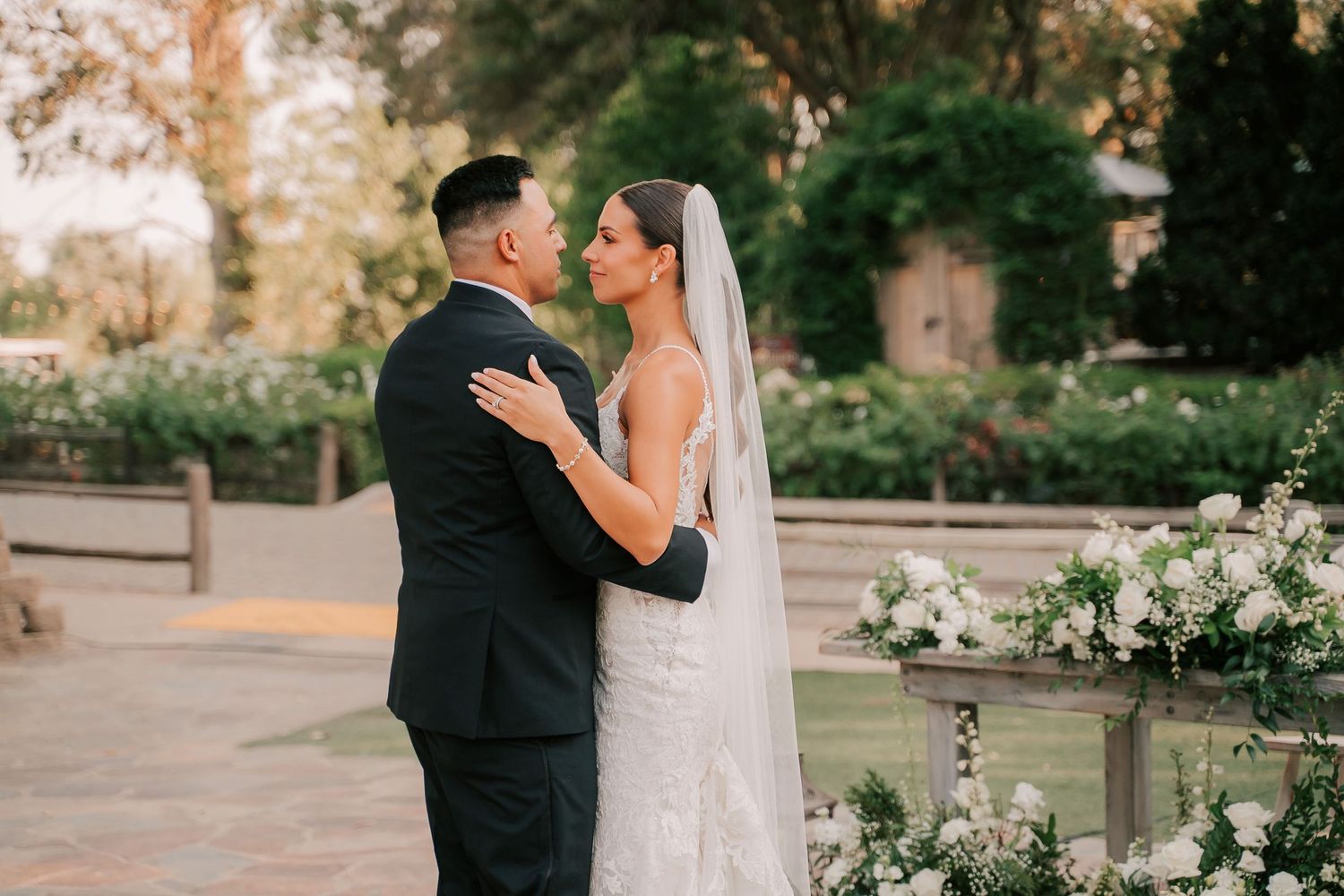 Bride and groom share a romantic embrace outdoors at a garden wedding ceremony with white floral arrangements.