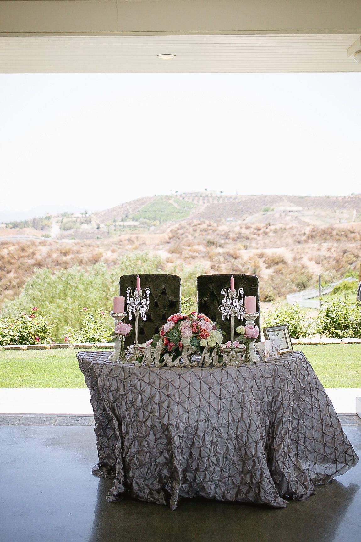 Elegant wedding sweetheart table with gray pintuck linen, crystal candelabras, pink floral centerpiece, and Mr. & Mrs. sign outdoors.