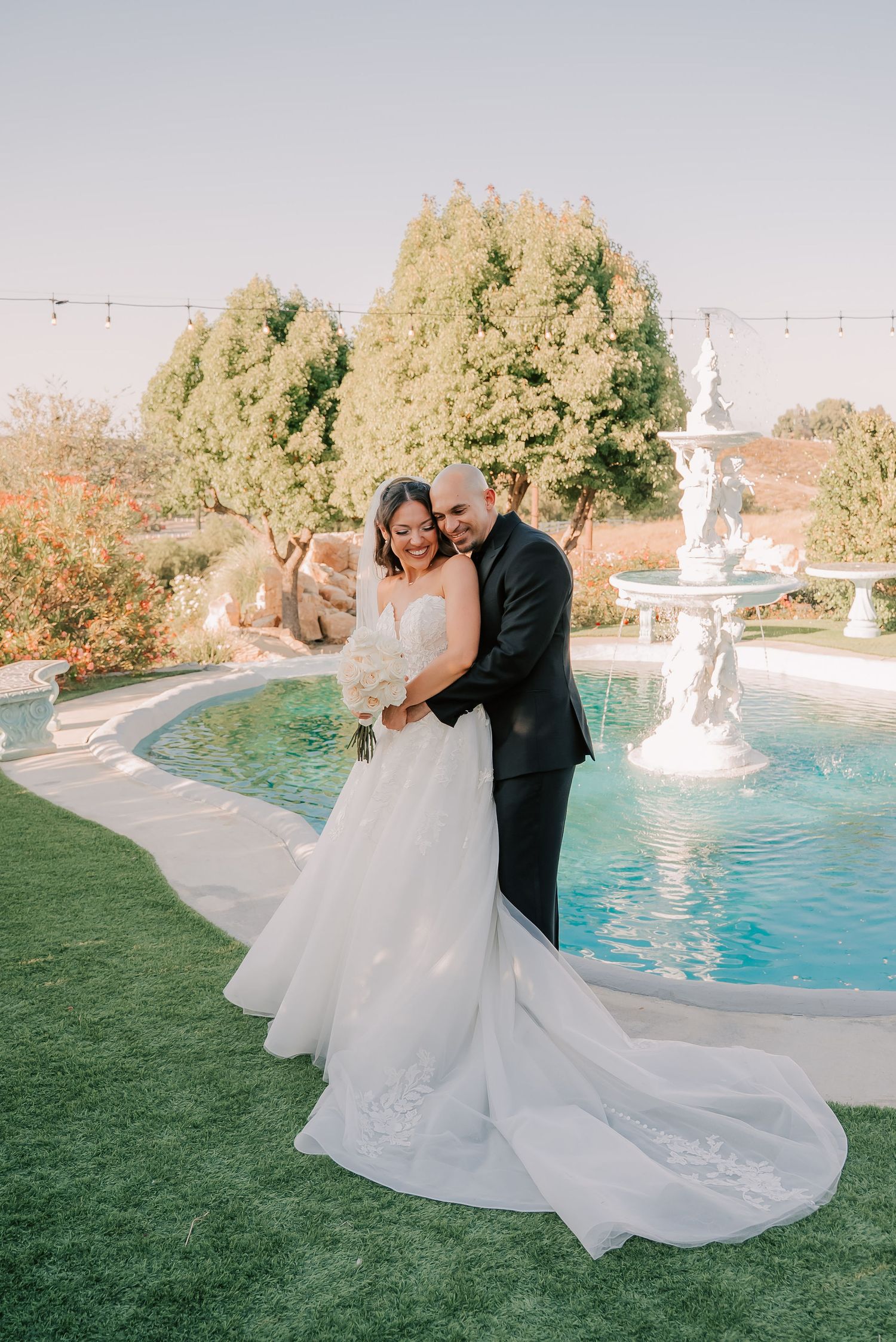 Bride and groom embrace near elegant white fountain and pool at outdoor wedding venue with string lights.