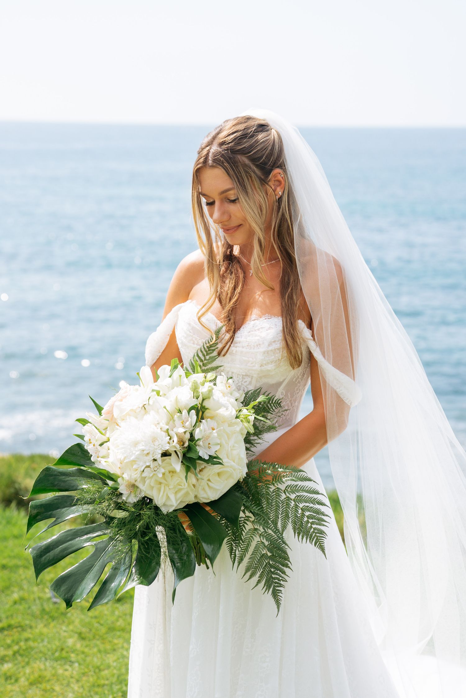 Bride in white off-shoulder gown and veil holds tropical bouquet of white flowers and ferns by the ocean.