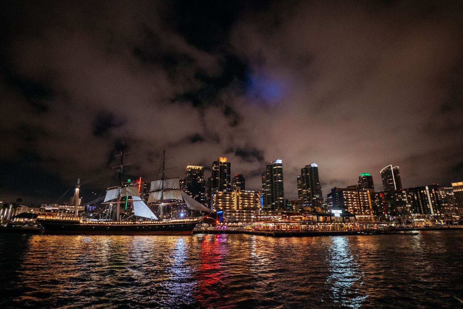 Tall ship and city skyline reflecting on water at night with dramatic cloudy sky in San Diego harbor.