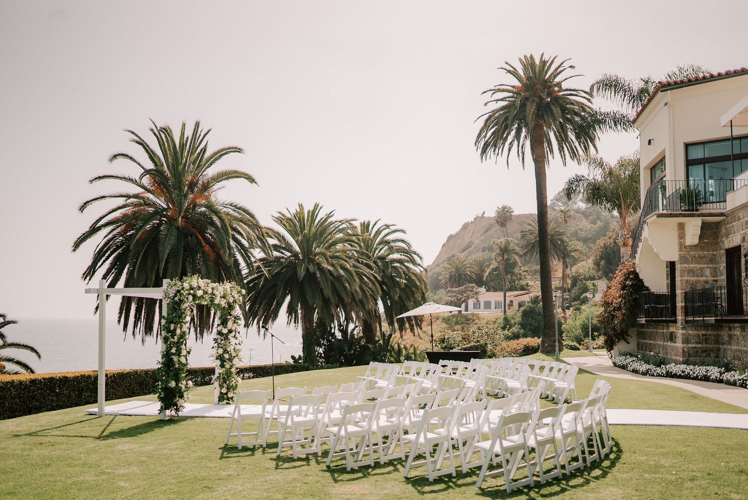 Outdoor wedding ceremony setup with white chairs and a floral arch on a manicured lawn, framed by palm trees and an oceanfront hillside.