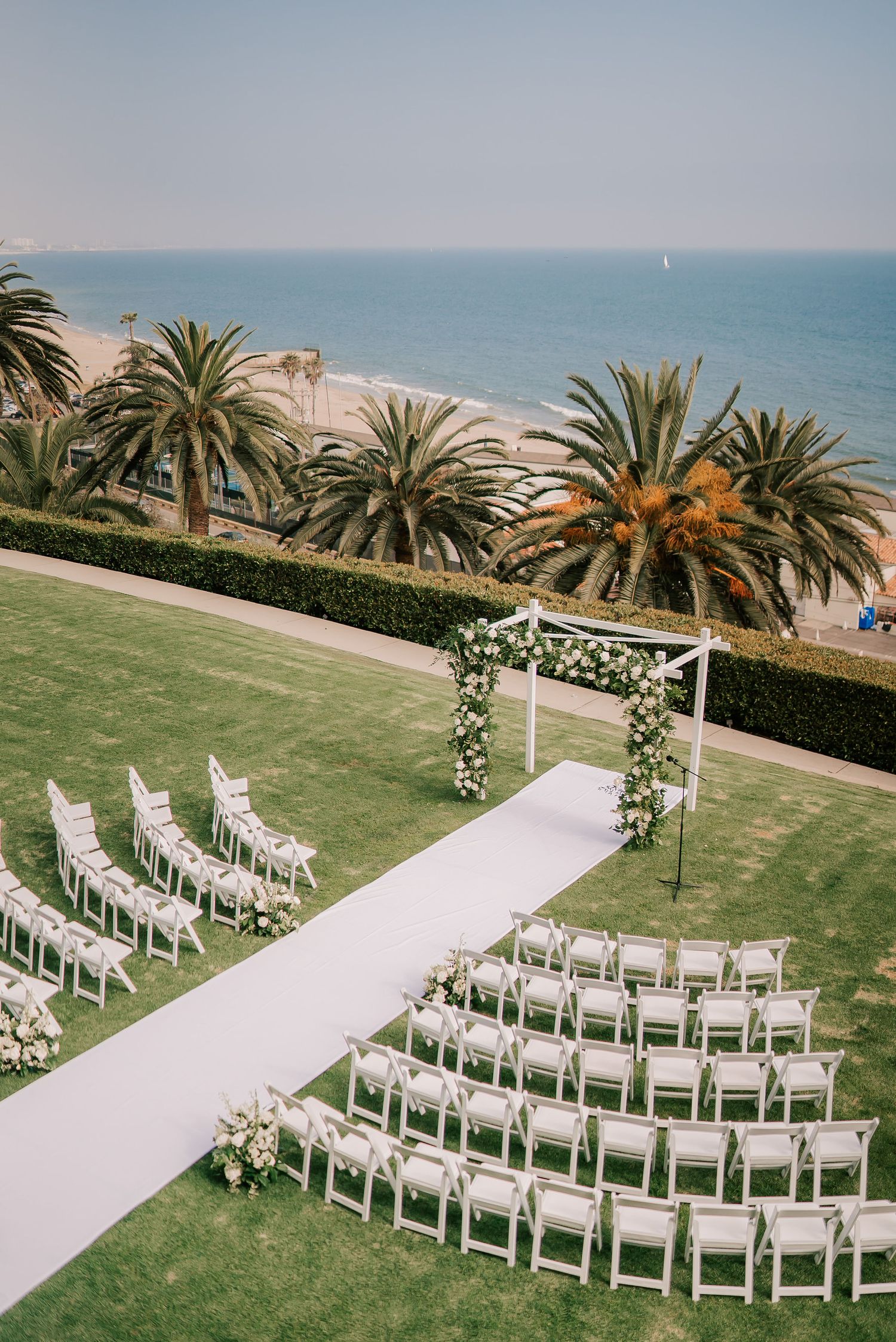 Oceanfront wedding ceremony setup with white chairs, a white aisle runner, and a floral arch on a green lawn lined with palm trees.