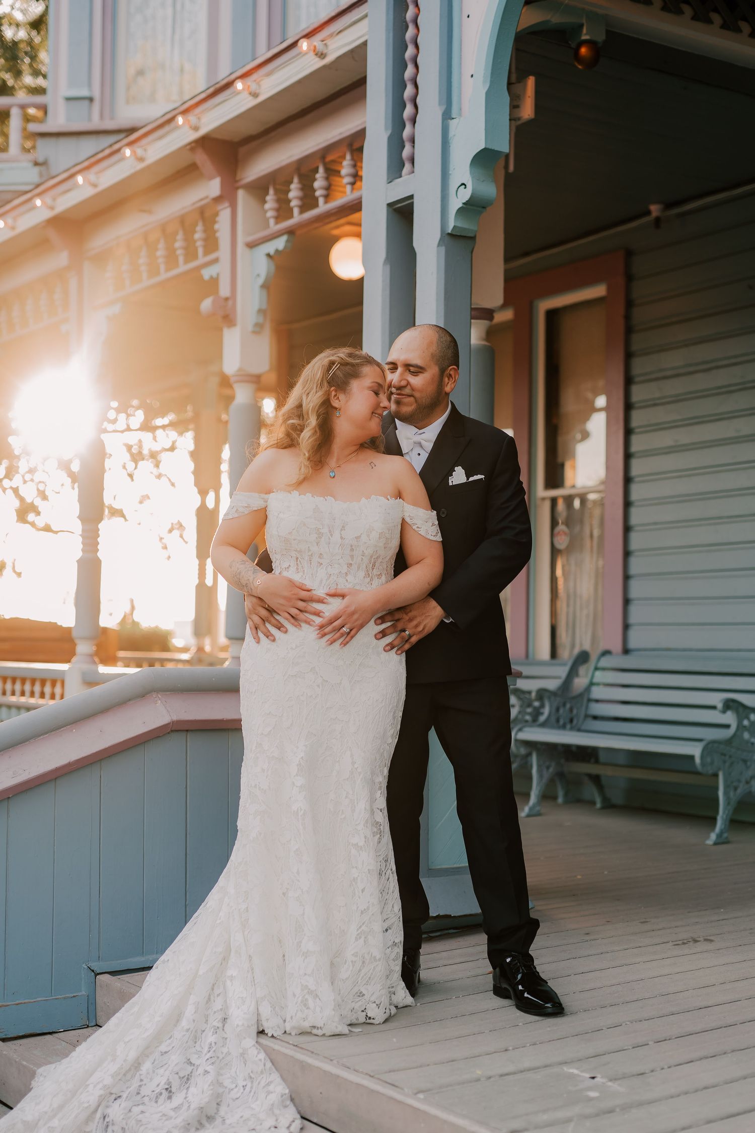 Bride and groom share a romantic moment on a Victorian porch at golden hour, bride in lace off-shoulder gown.
