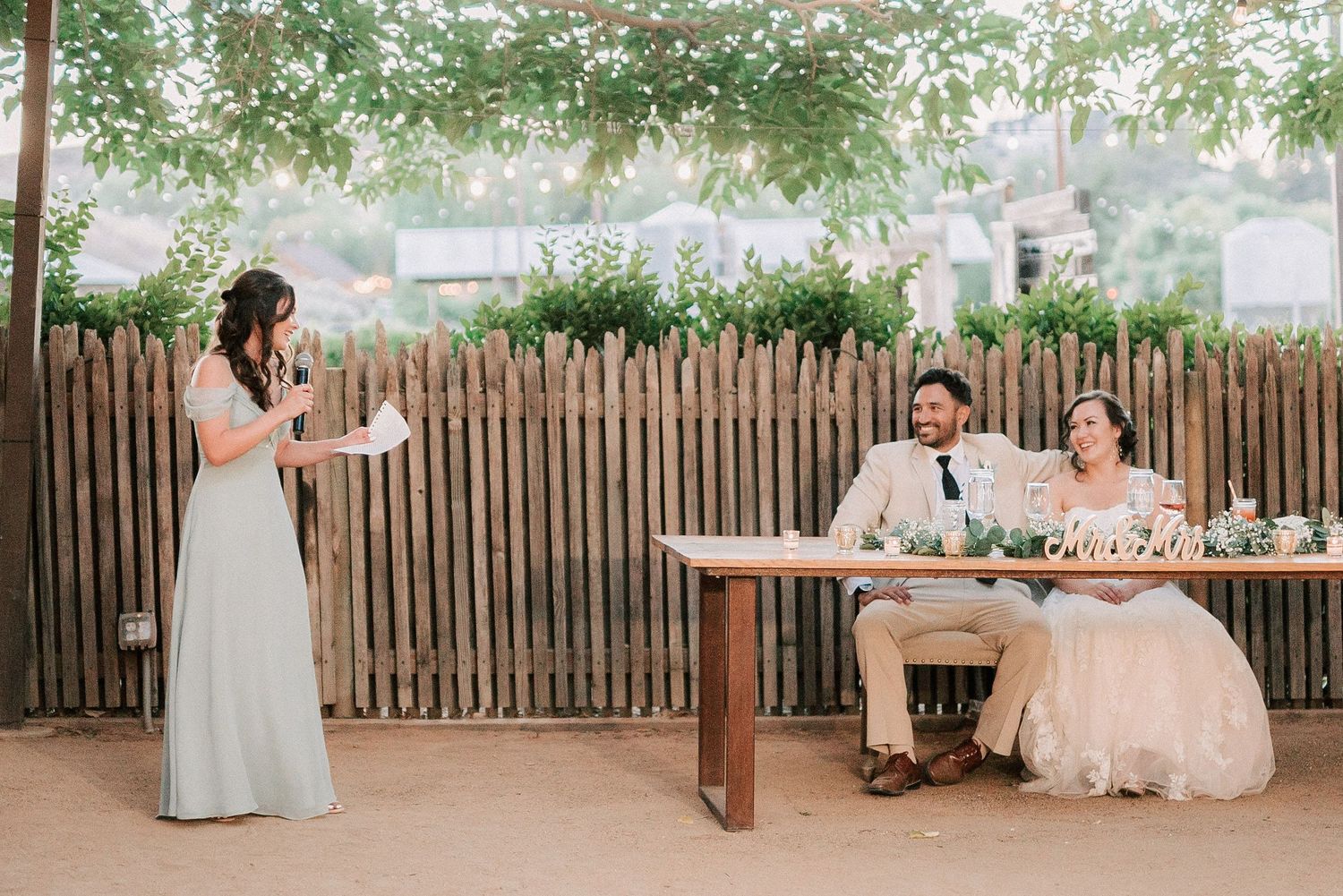 Bridesmaid giving a toast speech at an outdoor wedding reception while bride and groom smile at sweetheart table.