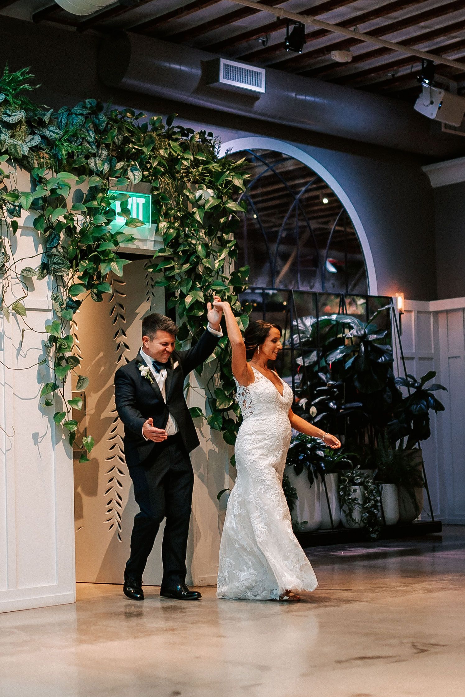 Bride and groom share first dance under lush greenery arch at elegant indoor wedding venue.