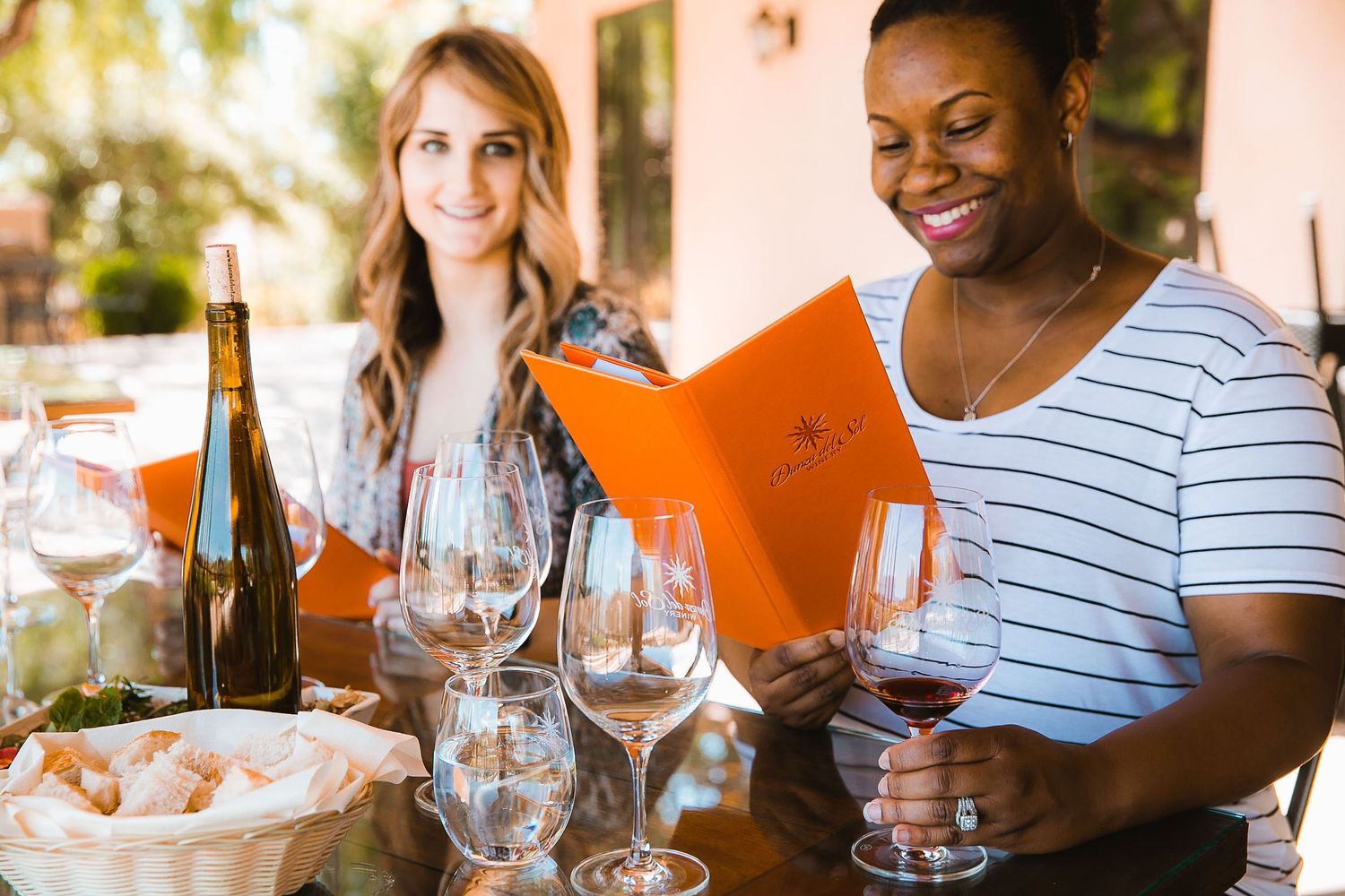 Two women smiling and reviewing a menu with wine glasses at an outdoor restaurant.