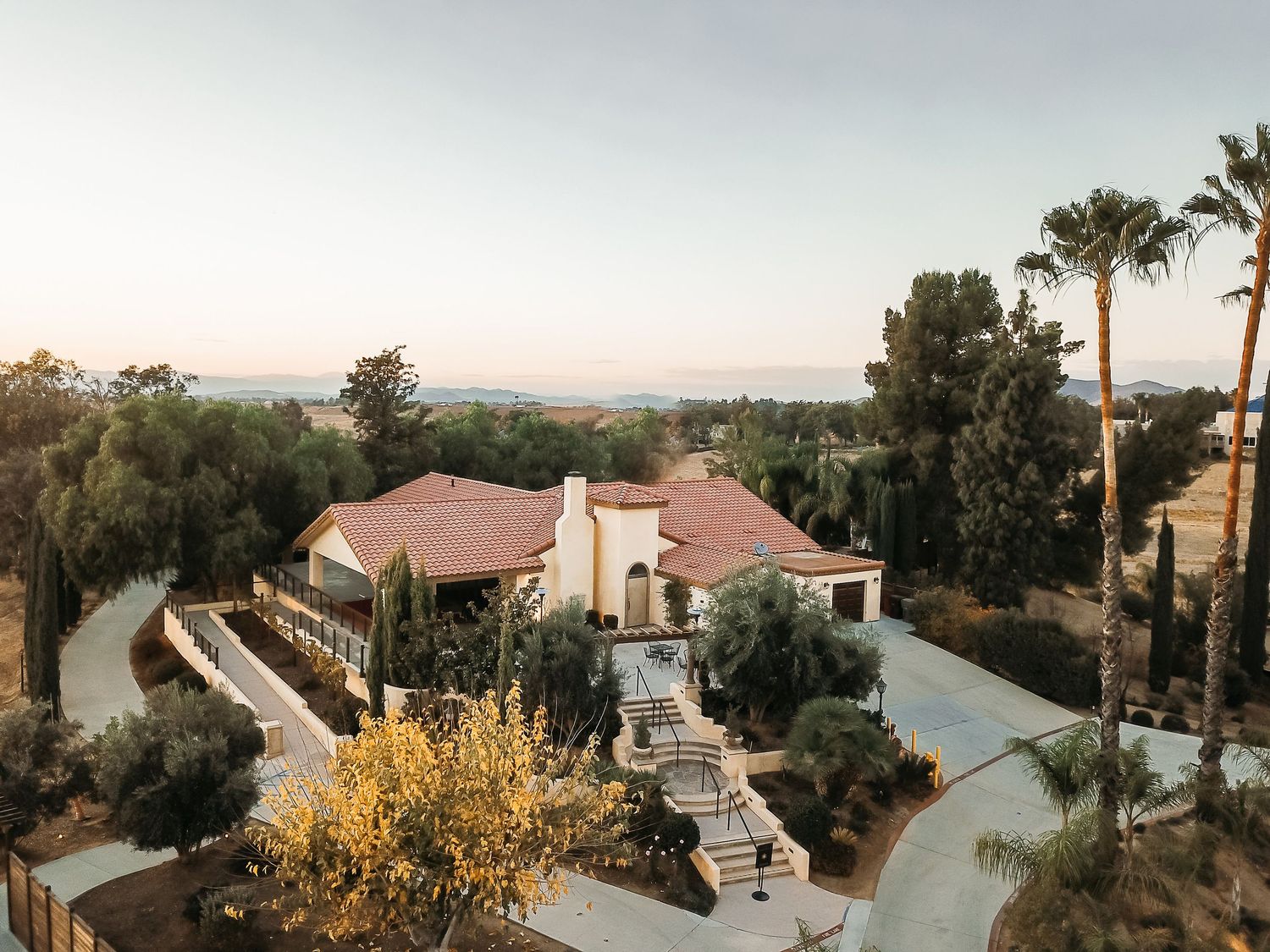 Aerial view of a luxurious Mediterranean-style estate surrounded by lush trees with panoramic valley views at dusk.