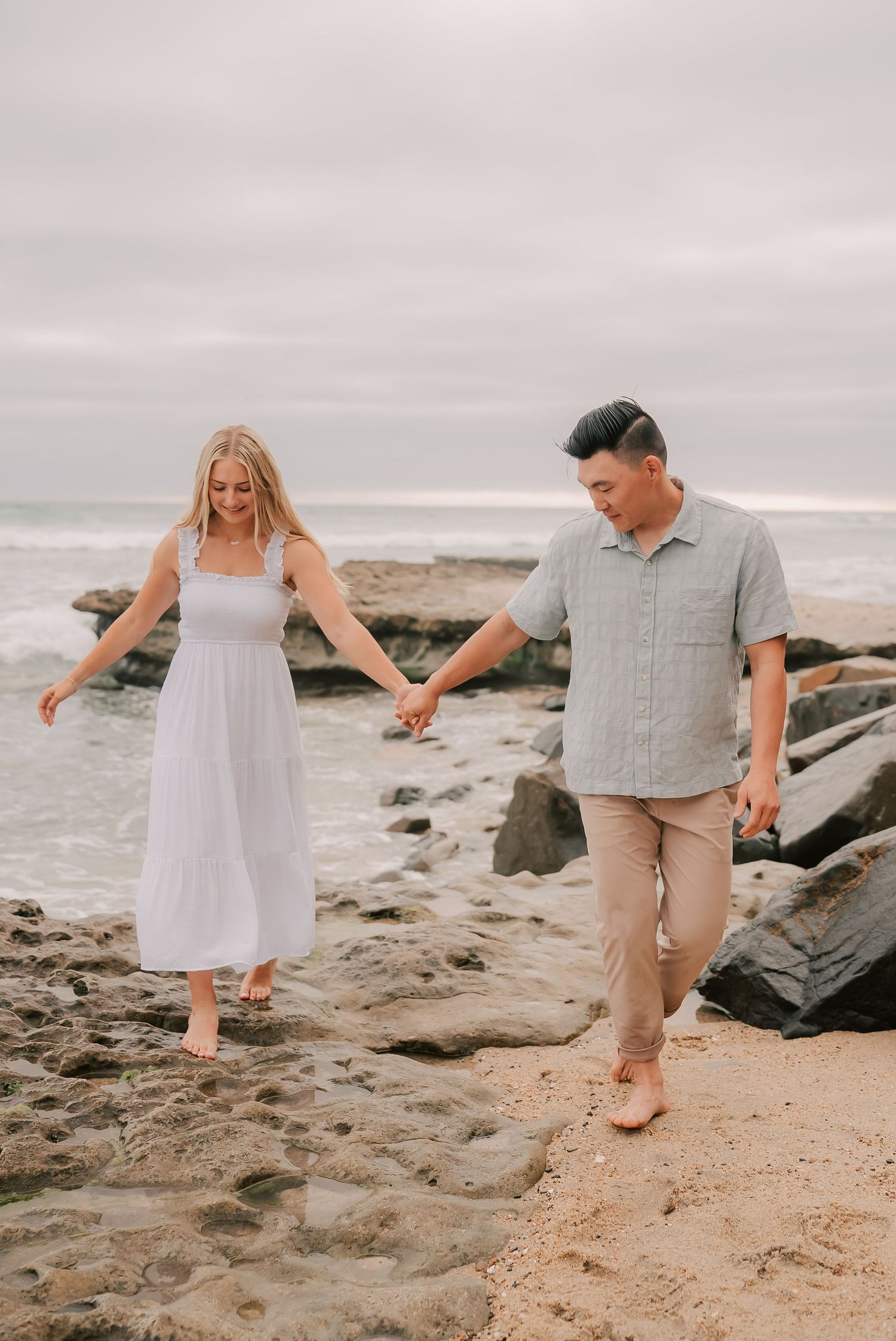 Couple walking hand-in-hand barefoot on rocky beach, woman in white dress, man in gray shirt and tan pants.