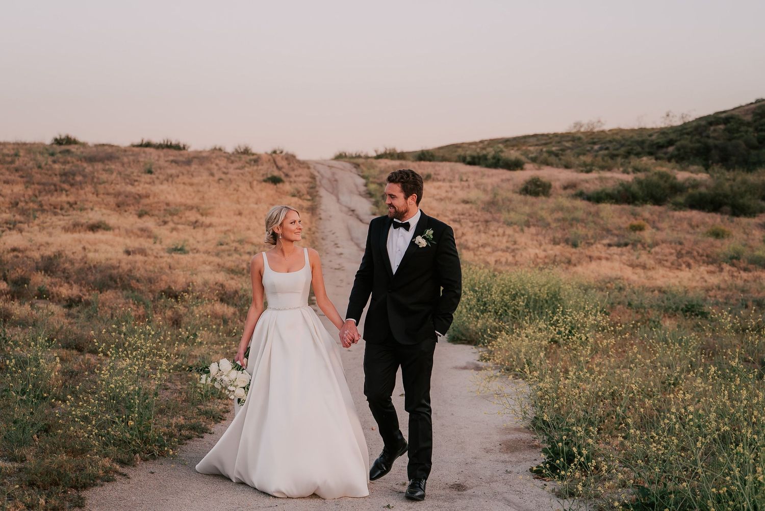 Bride and groom walk hand-in-hand along a scenic dirt path through golden fields at sunset.