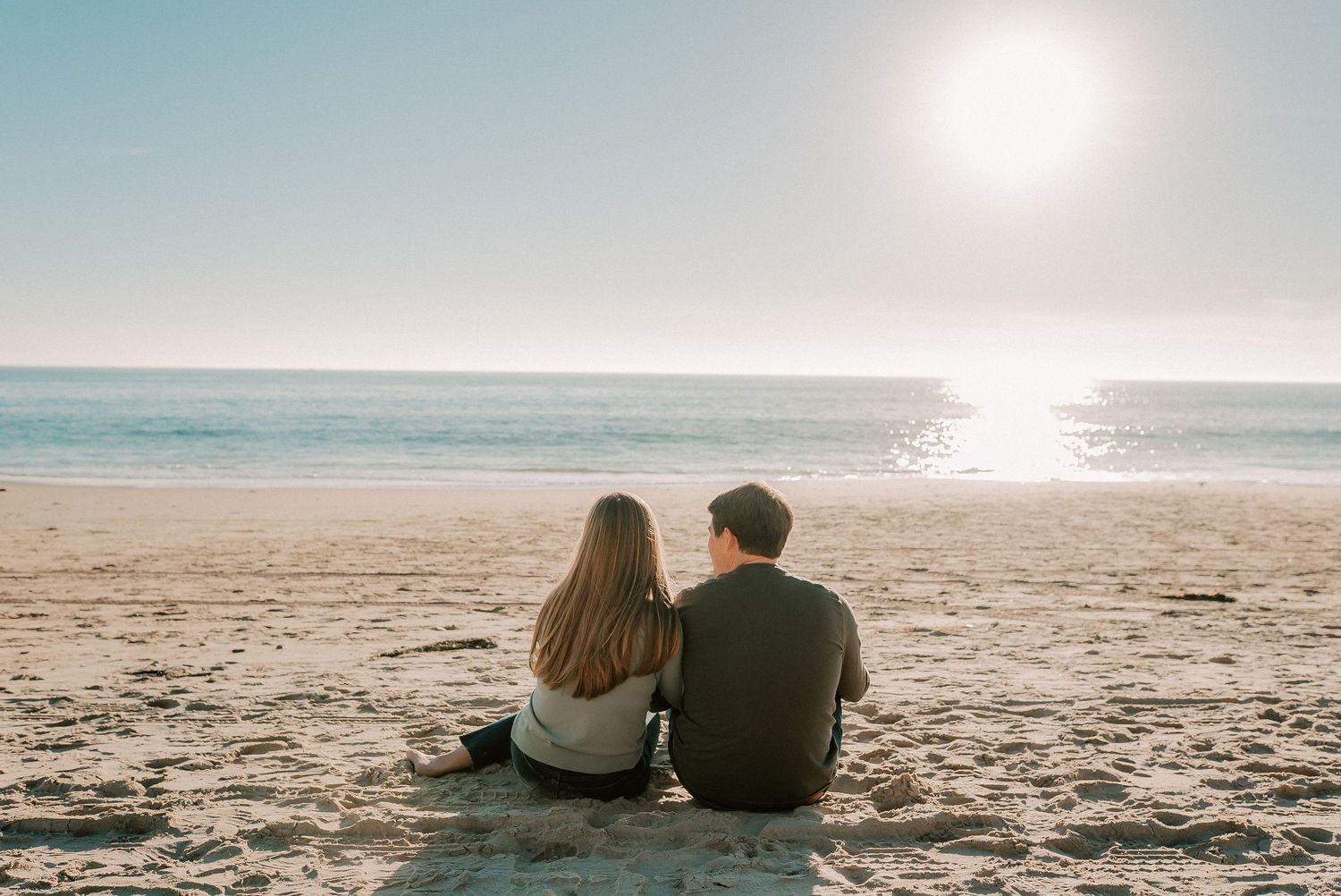 Couple sitting together on sandy beach watching sunset over calm ocean waves in romantic seaside setting.