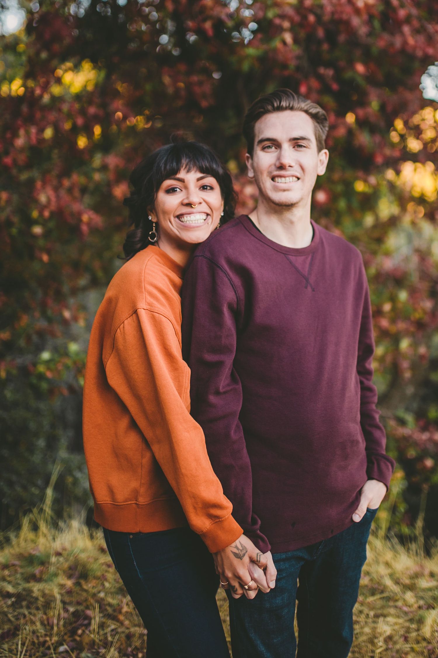 A smiling couple holds hands outdoors in fall, wearing orange and burgundy sweatshirts with colorful autumn foliage behind them.