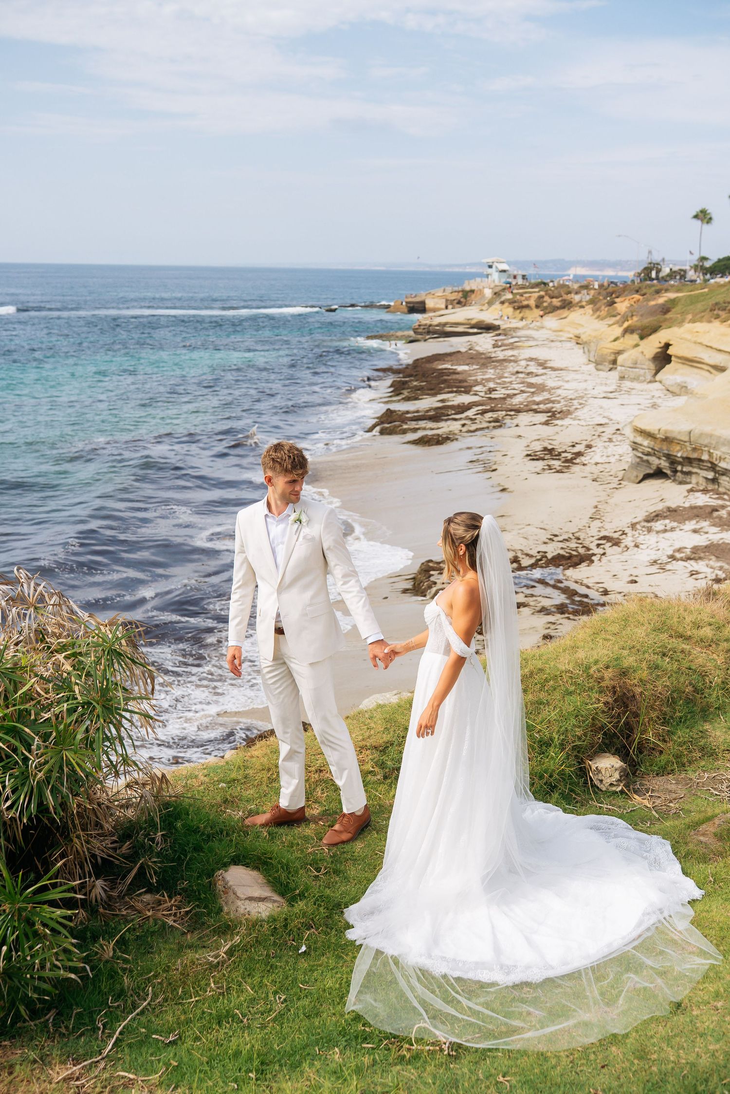 Bride and groom hold hands on coastal cliffside, her white gown trailing behind with ocean views.