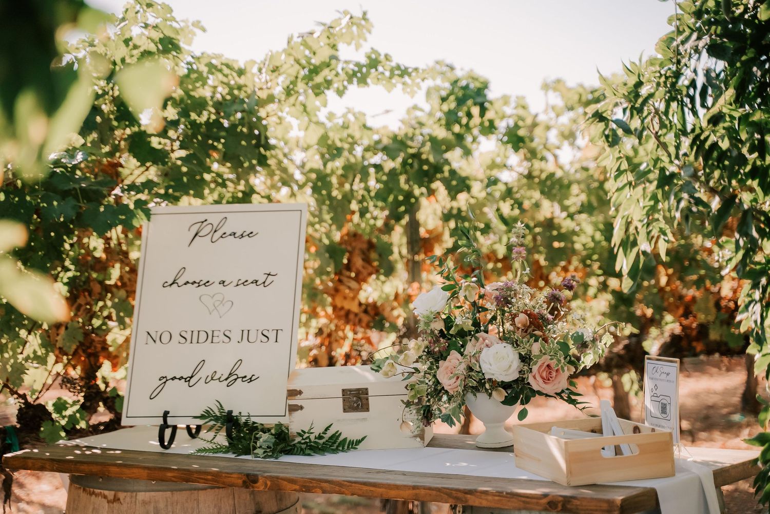 Rustic wedding seating sign reading 'No Sides Just Good Vibes' with blush floral arrangement on wooden table outdoors.