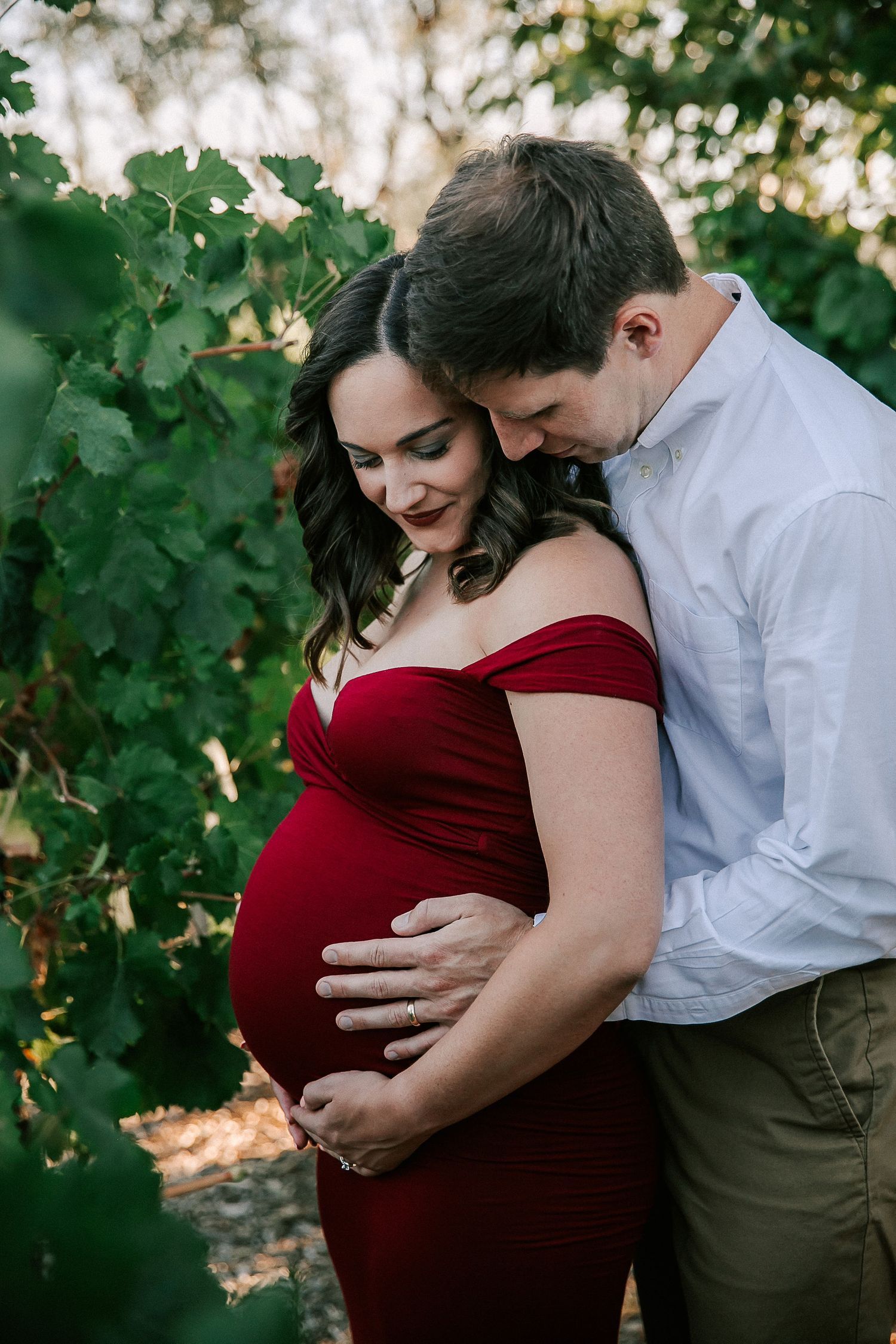 Pregnant woman in red off-shoulder gown embraced by partner among lush green vineyard leaves during maternity photo session.