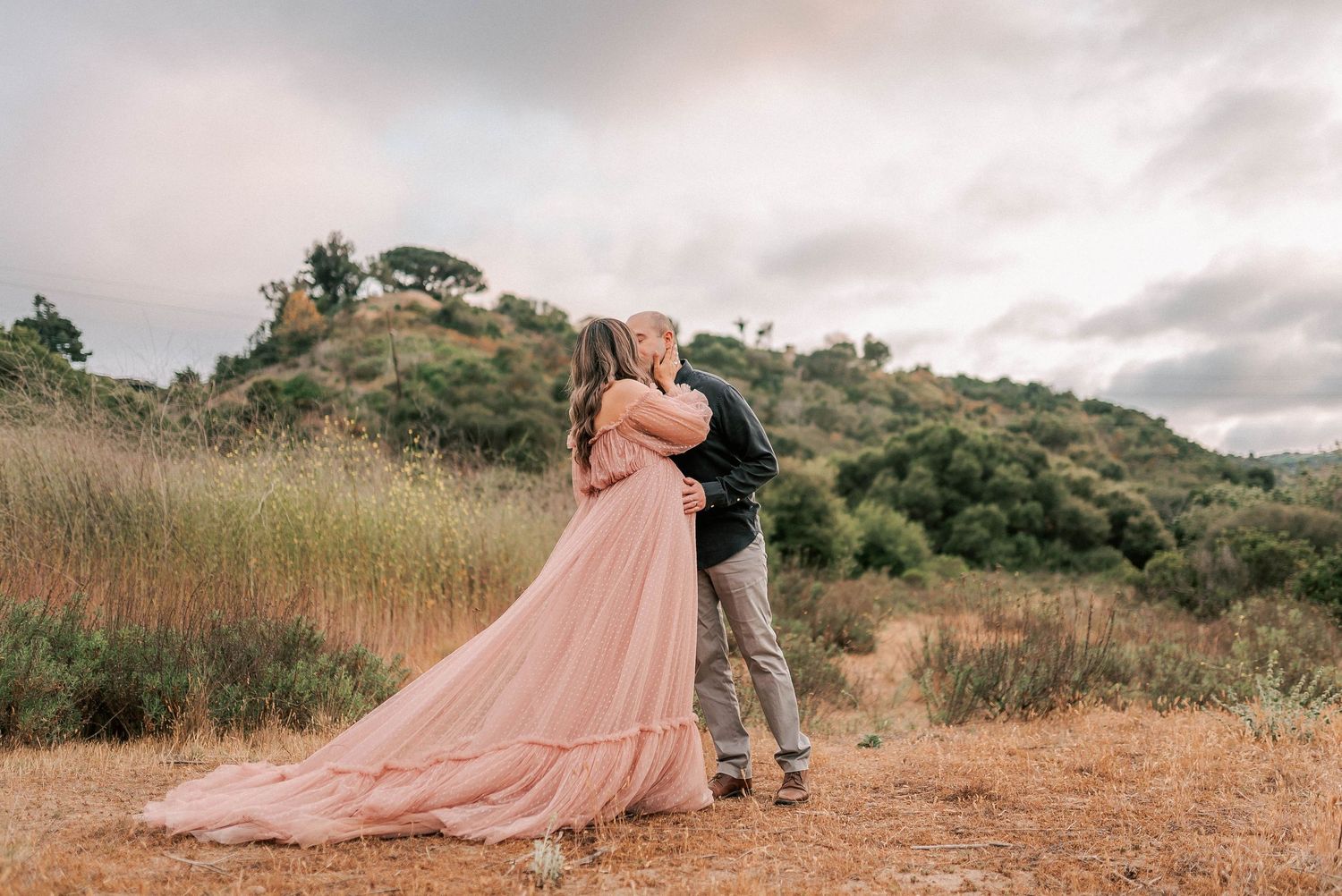 Couple embracing in a field, woman in flowing pink gown, lush green hills under cloudy sky.