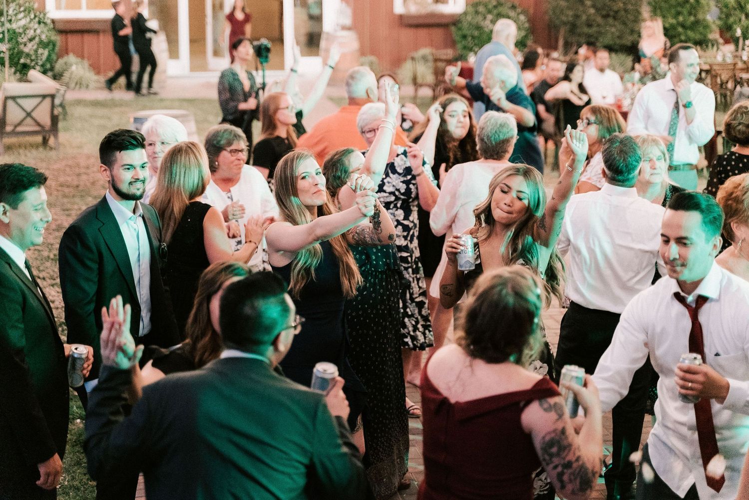 Guests dancing outdoors at a wedding reception, dressed in formal attire, enjoying the celebration under green ambient lighting.