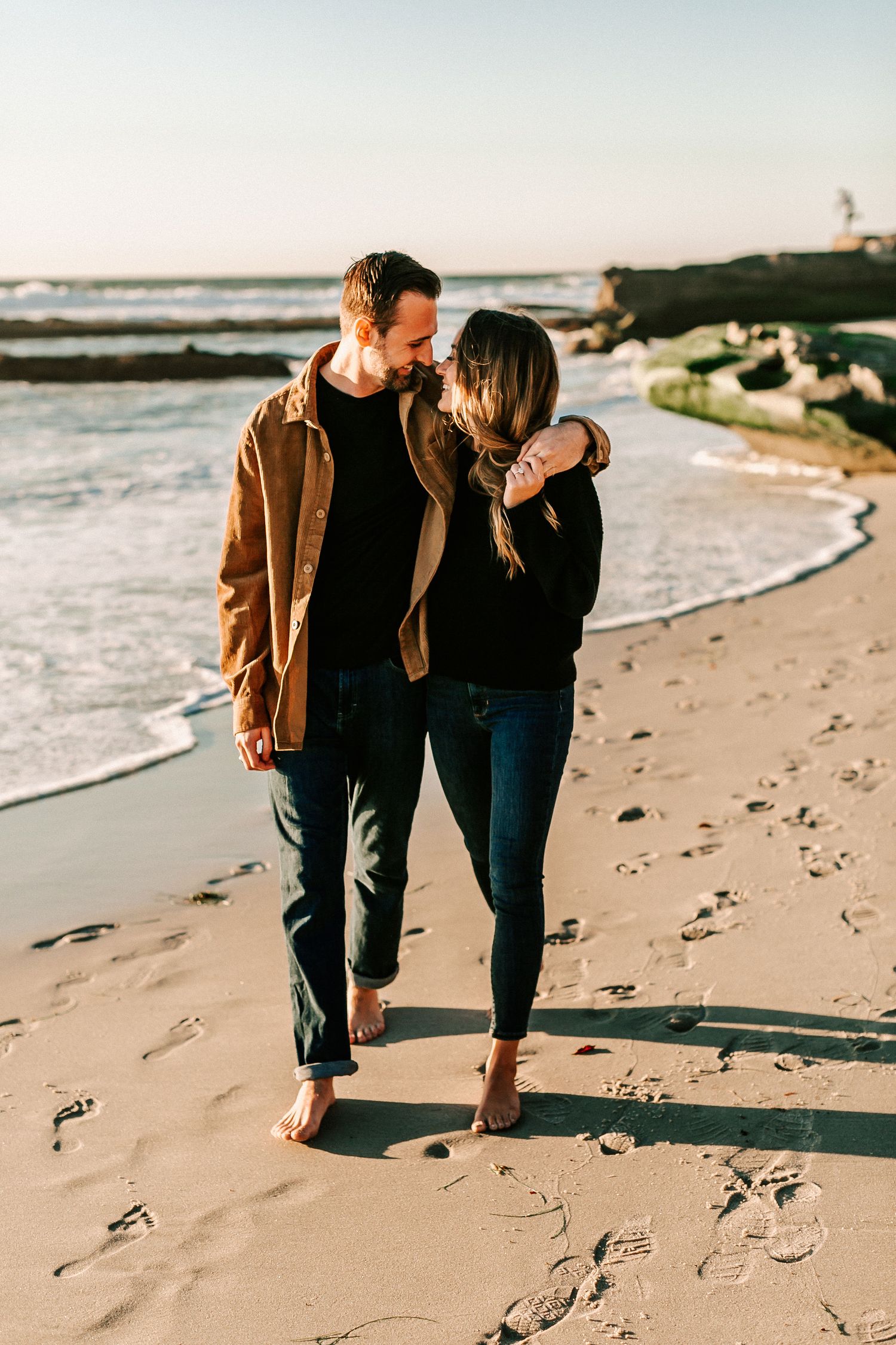 Barefoot couple walking arm in arm along sandy beach at golden sunset.