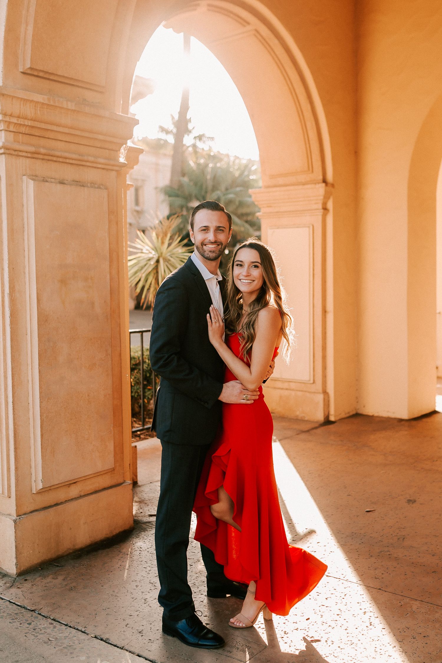 Couple embracing under a sunlit archway, woman in red dress and man in dark suit.