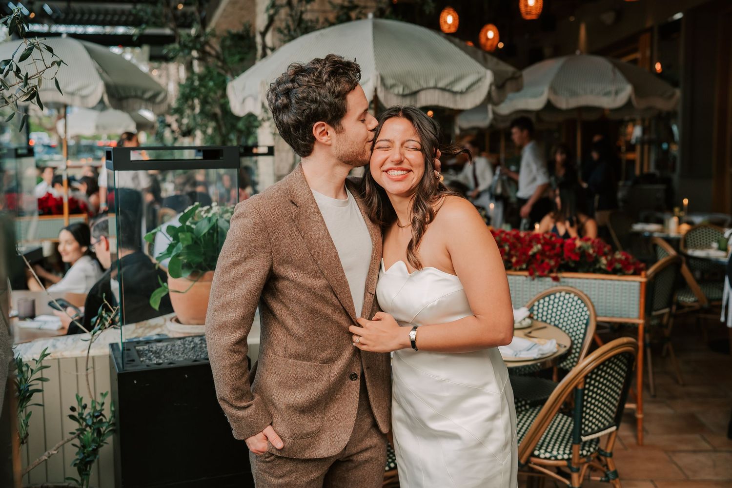 A groom in a brown suit kisses his smiling bride in a white dress at an outdoor restaurant wedding reception.