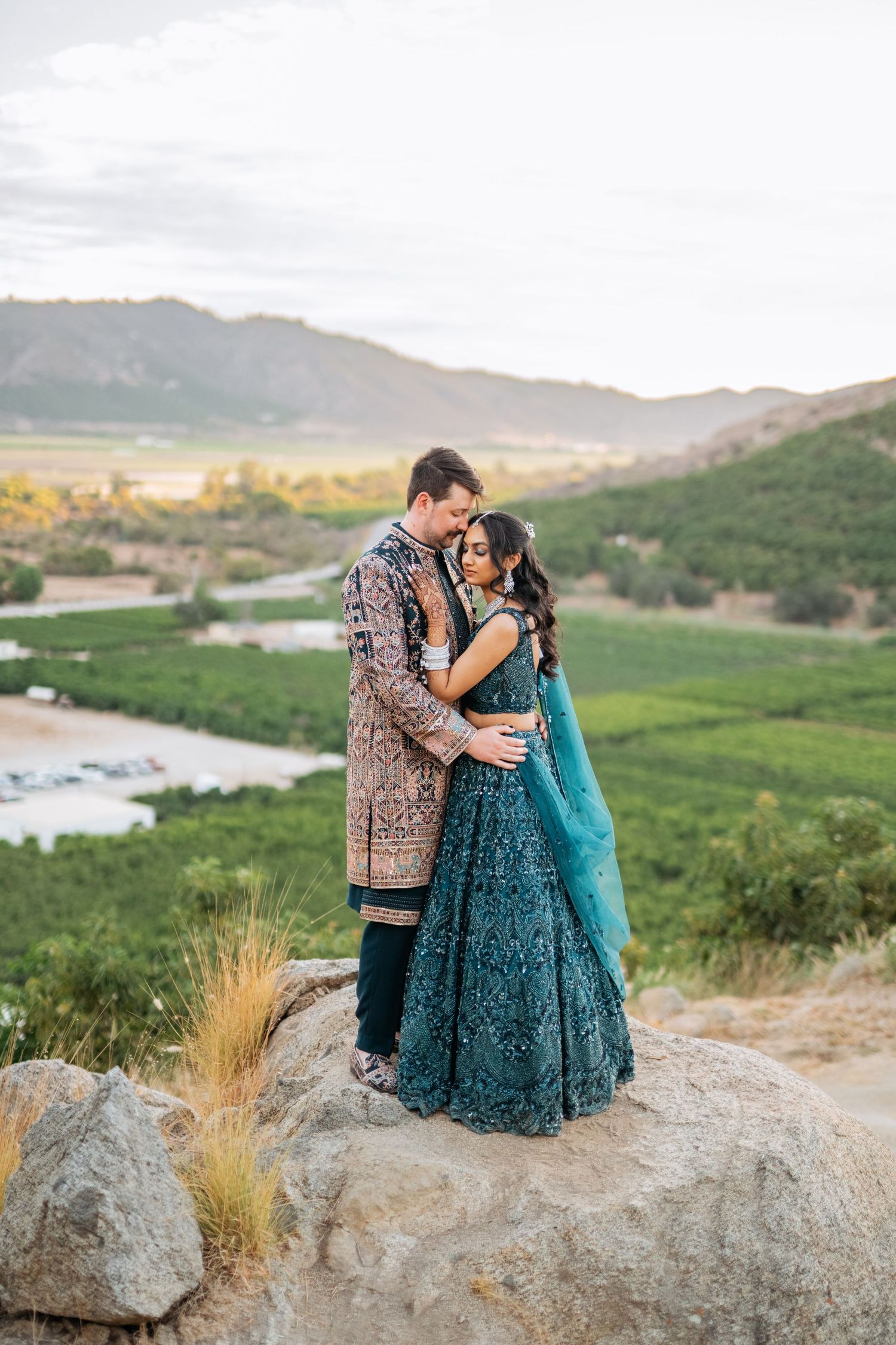 Couple in Indian wedding attire embracing on rocks with scenic vineyard and mountain backdrop at sunset.
