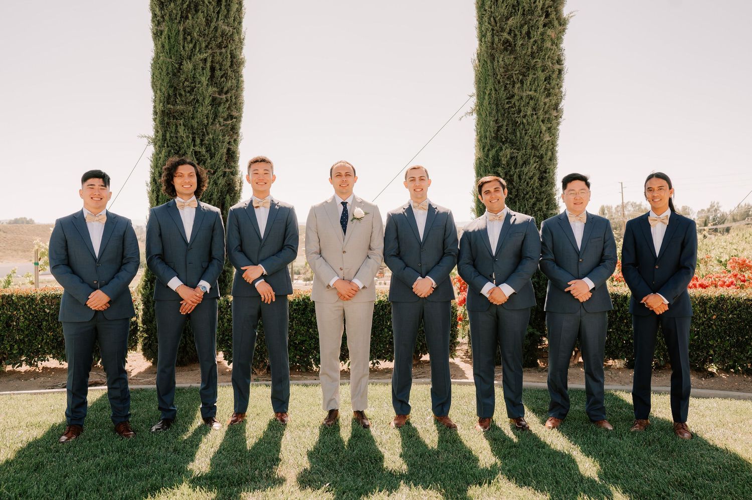 Groom in tan suit stands with eight groomsmen in navy suits outdoors at a wedding venue with cypress trees.