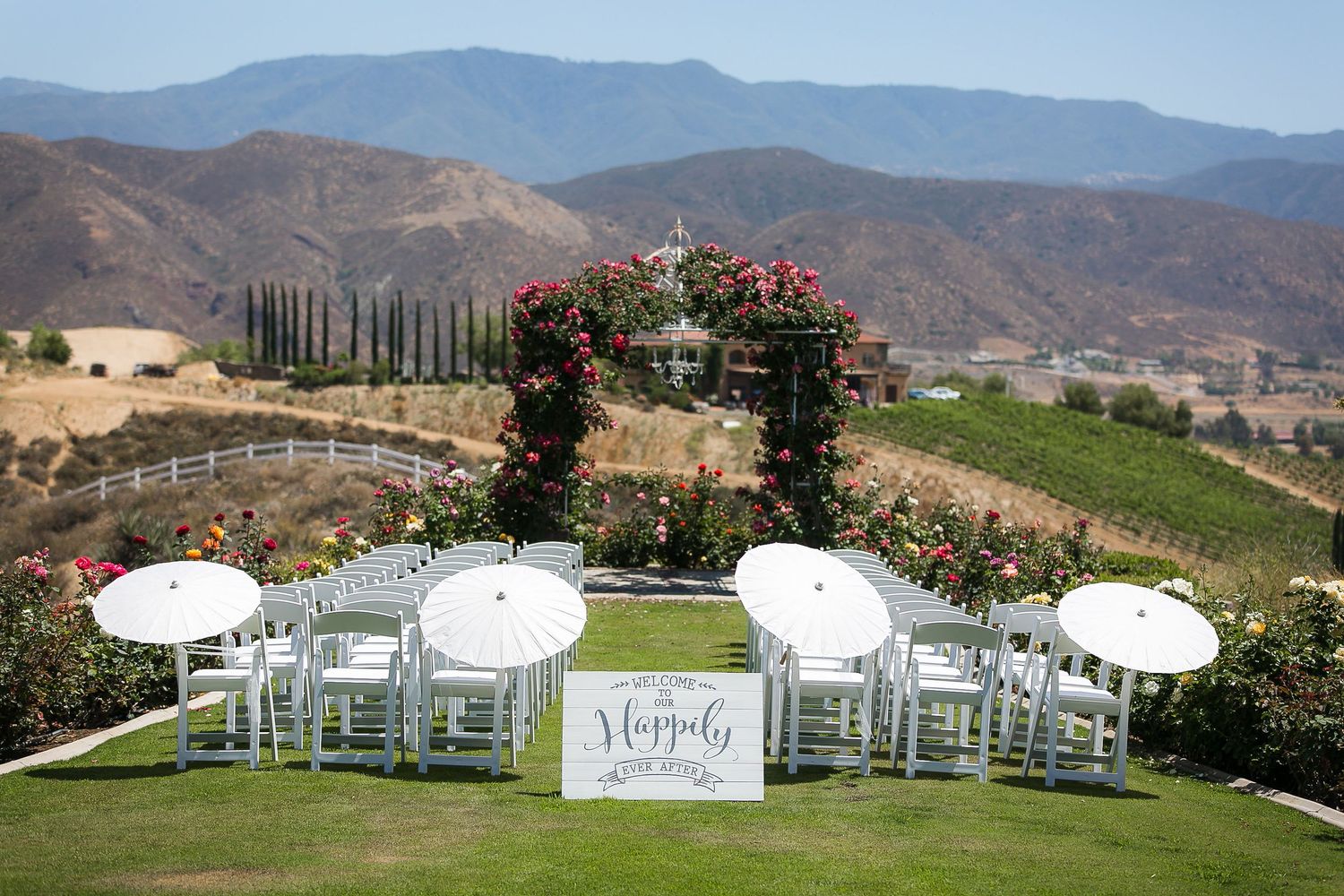 Outdoor vineyard wedding ceremony setup with white chairs, floral arch, umbrellas, and mountain views.