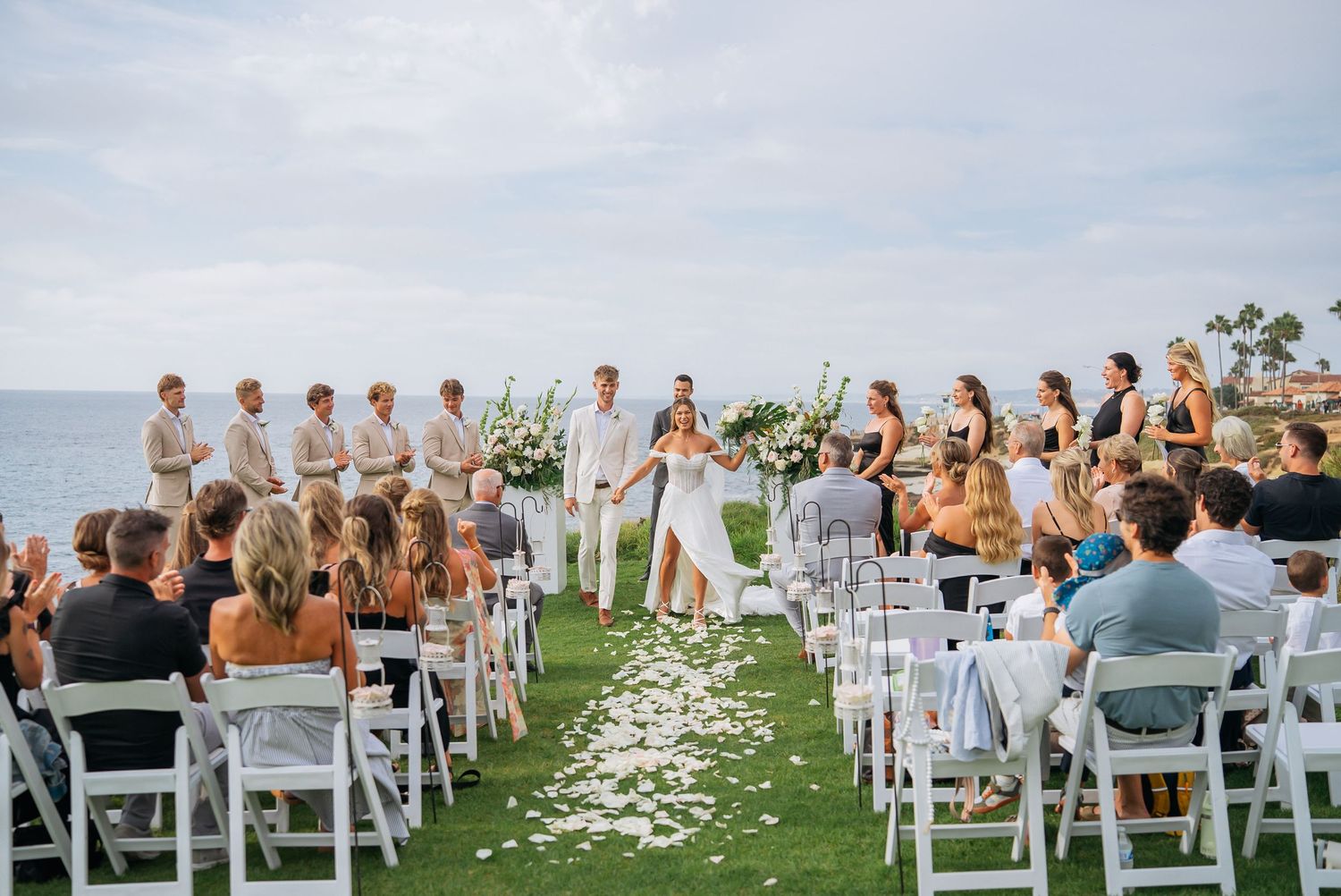 Aerial view of outdoor beach wedding ceremony with guests seated on white chairs along petal aisle.