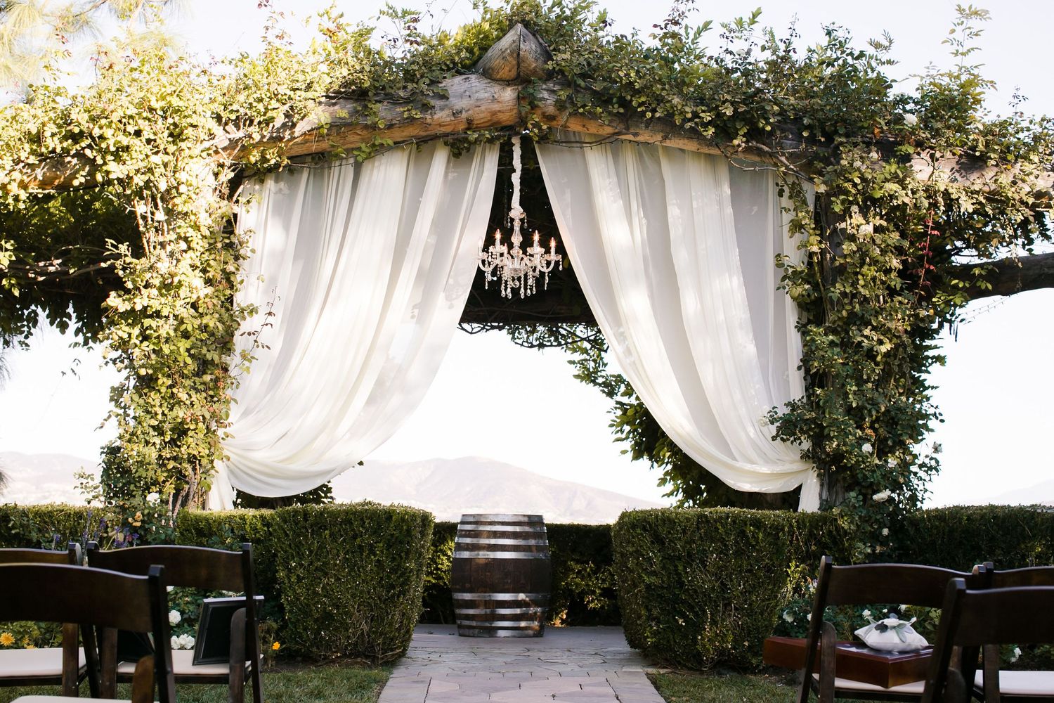 Elegant outdoor wedding ceremony arch adorned with white drapes, chandelier, and lush greenery, flanked by wooden barrel accents.