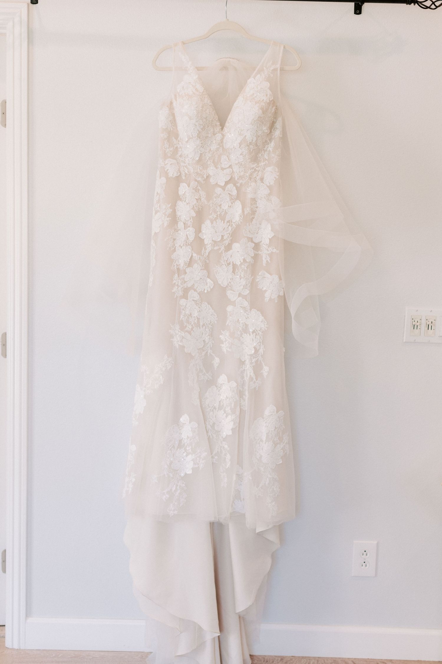 Elegant white lace floral wedding gown with V-neckline and tulle overlay hanging on a hanger against a white wall.