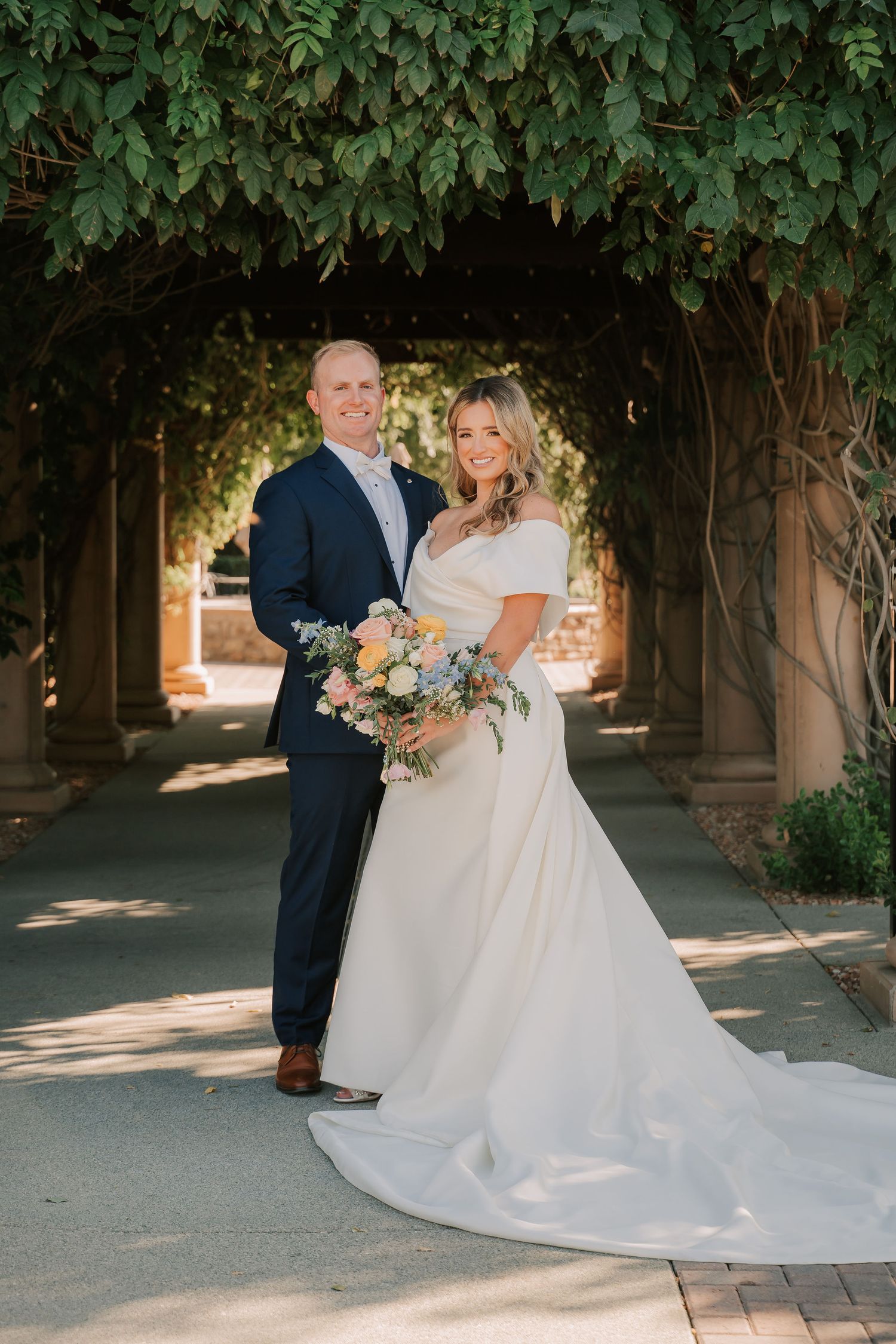 Couple in wedding attire pose under ivy-covered archway, bride holding pastel bouquet.