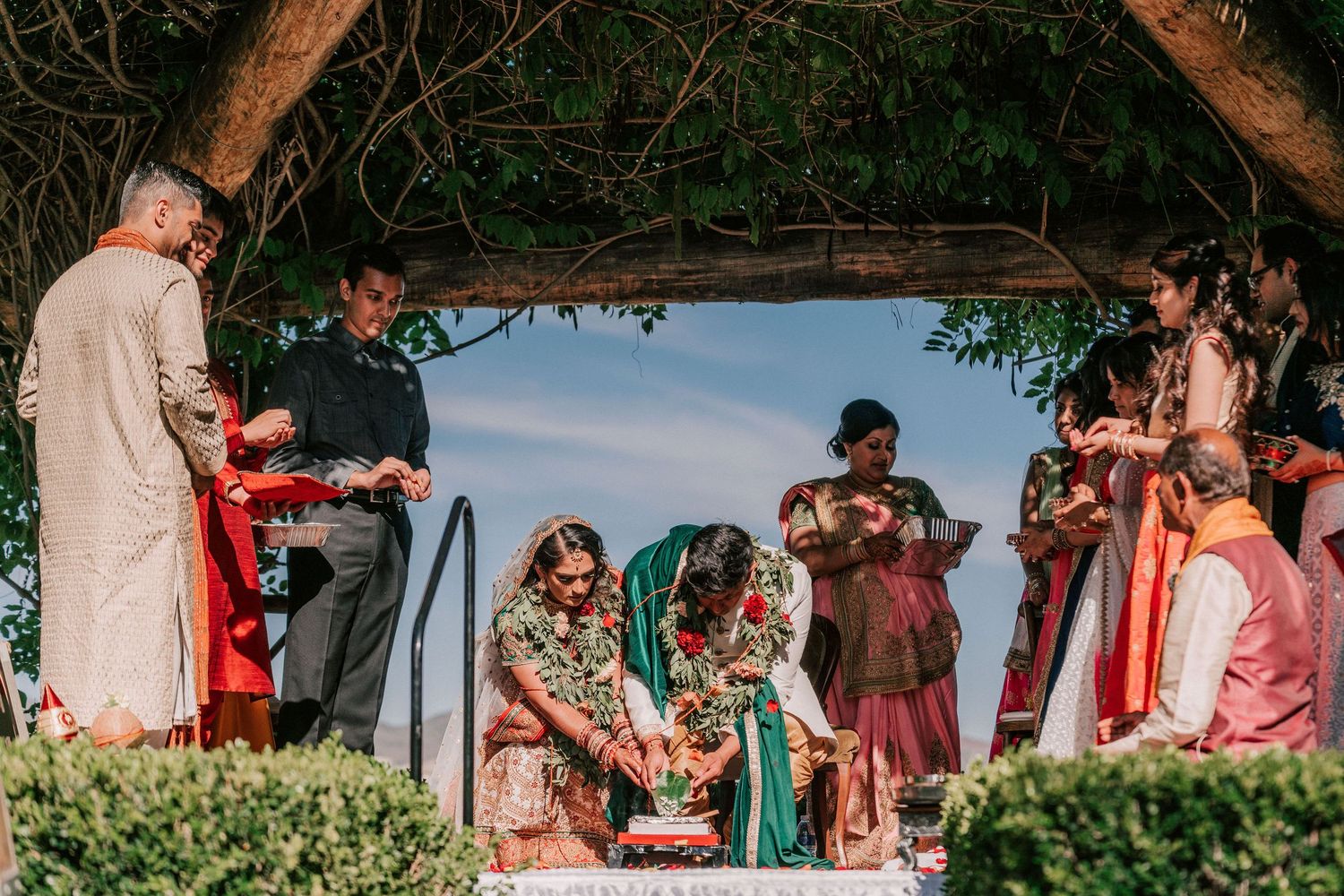 Indian wedding couple performing traditional rituals under pergola with family present.