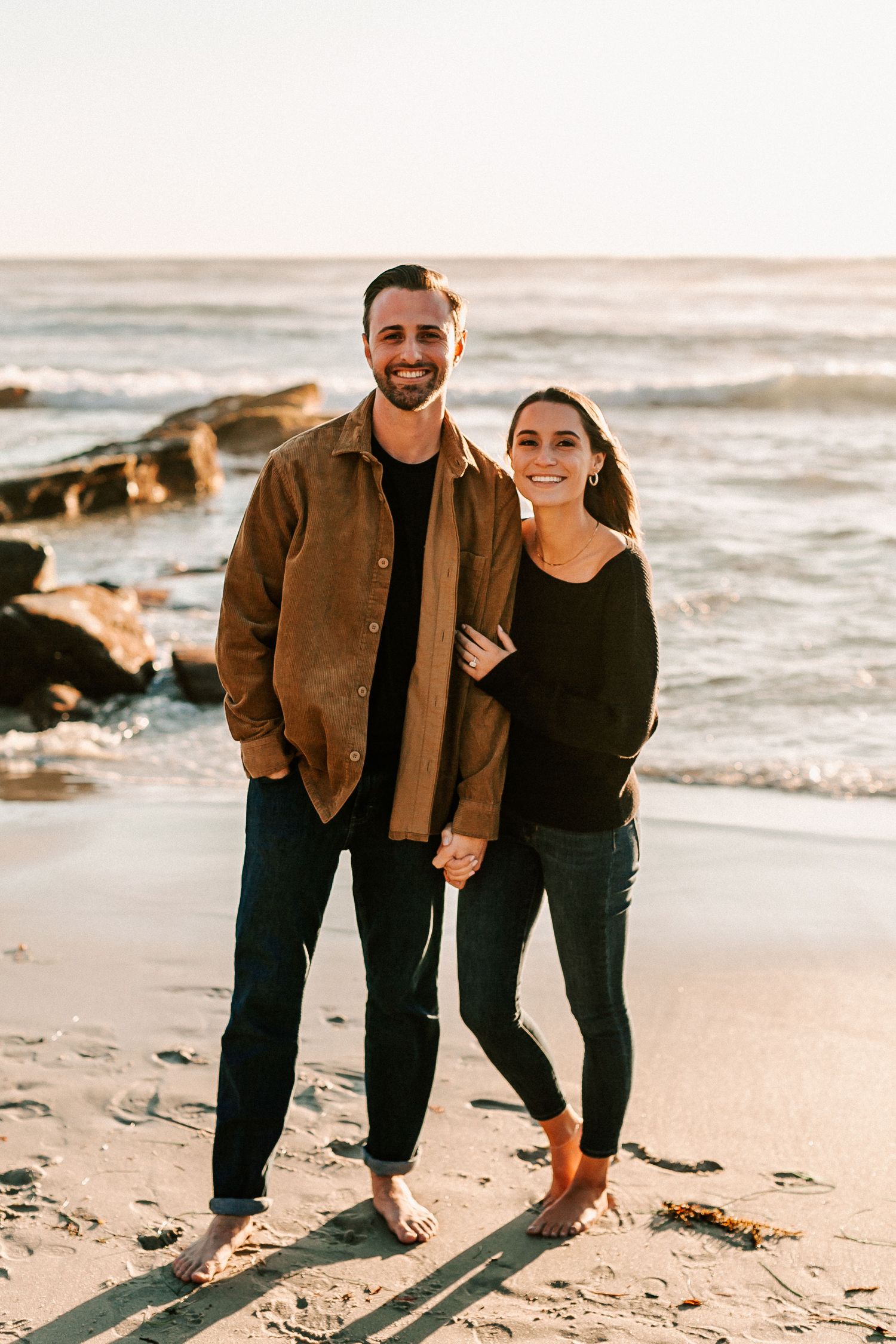 Couple smiling and holding hands on a sandy beach at golden hour with ocean waves in the background.