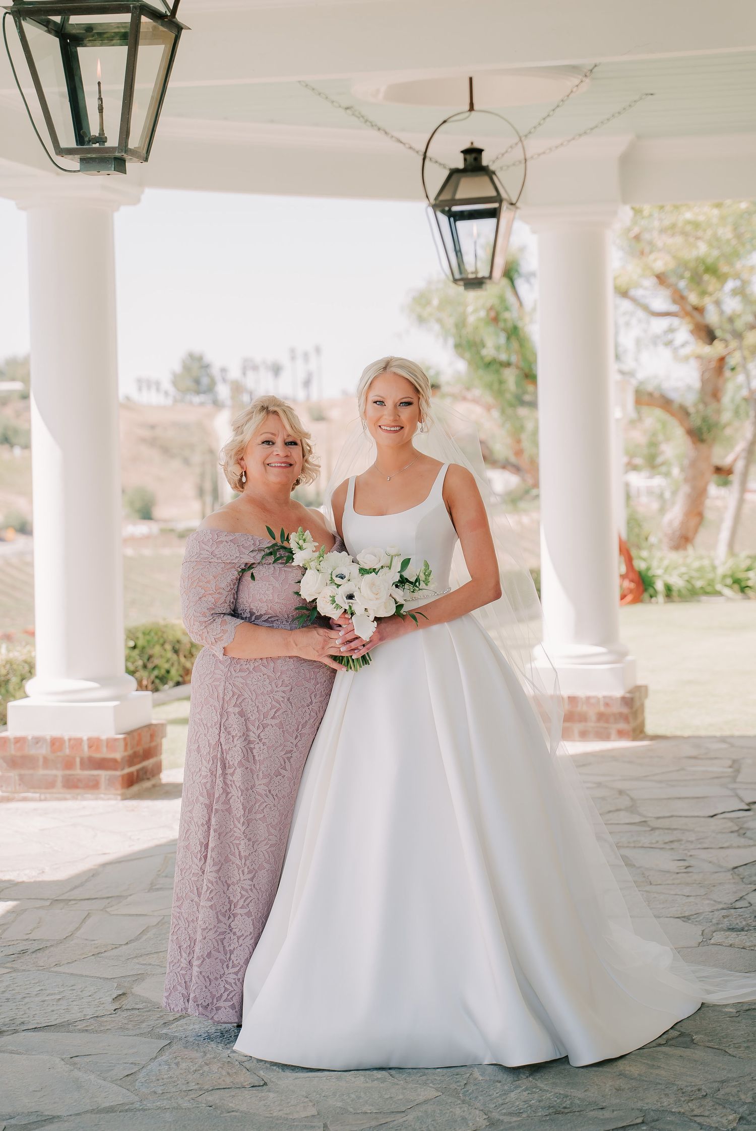 Bride in white square-neck ball gown poses with older woman in lavender lace dress under white columns.
