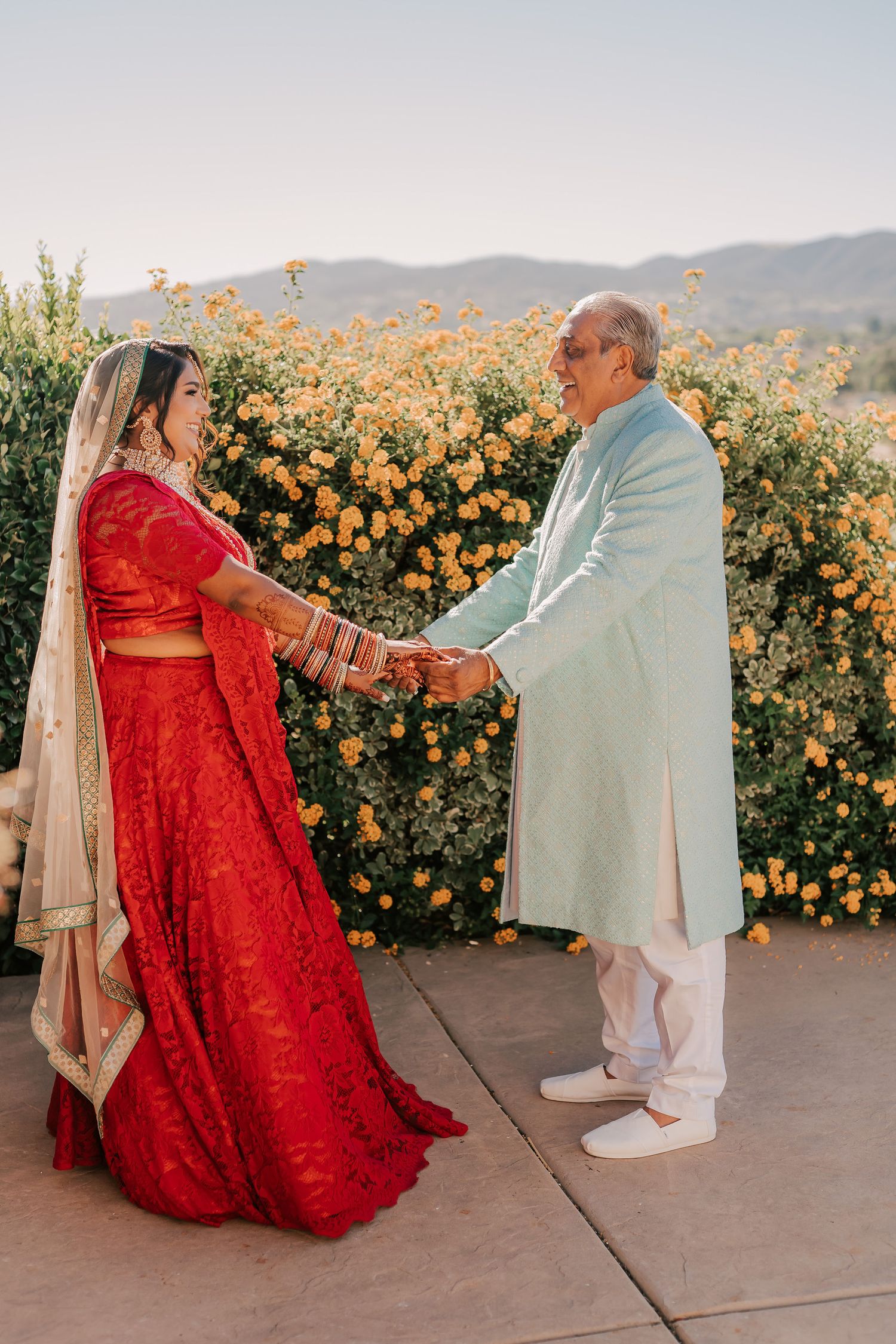 Indian bride in red lehenga holds hands with elderly man in light blue sherwani before yellow flower backdrop.