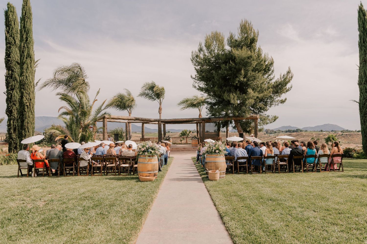 Wedding guests seated in rows on both sides of a grass aisle leading to a rustic wooden pergola at an outdoor vineyard ceremony.